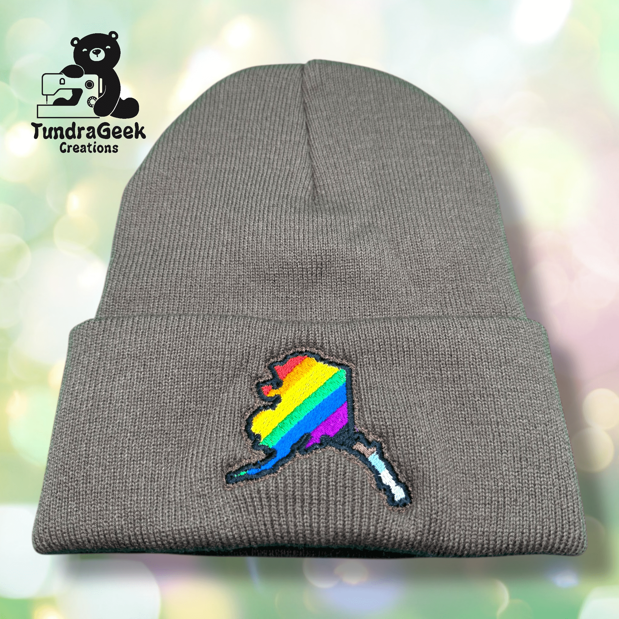 A gray knit beanie with an embroidered Alaska state outline in rainbow stripes, displayed on a blurred, colorful background. TundraGeek Creations logo in the top left corner.