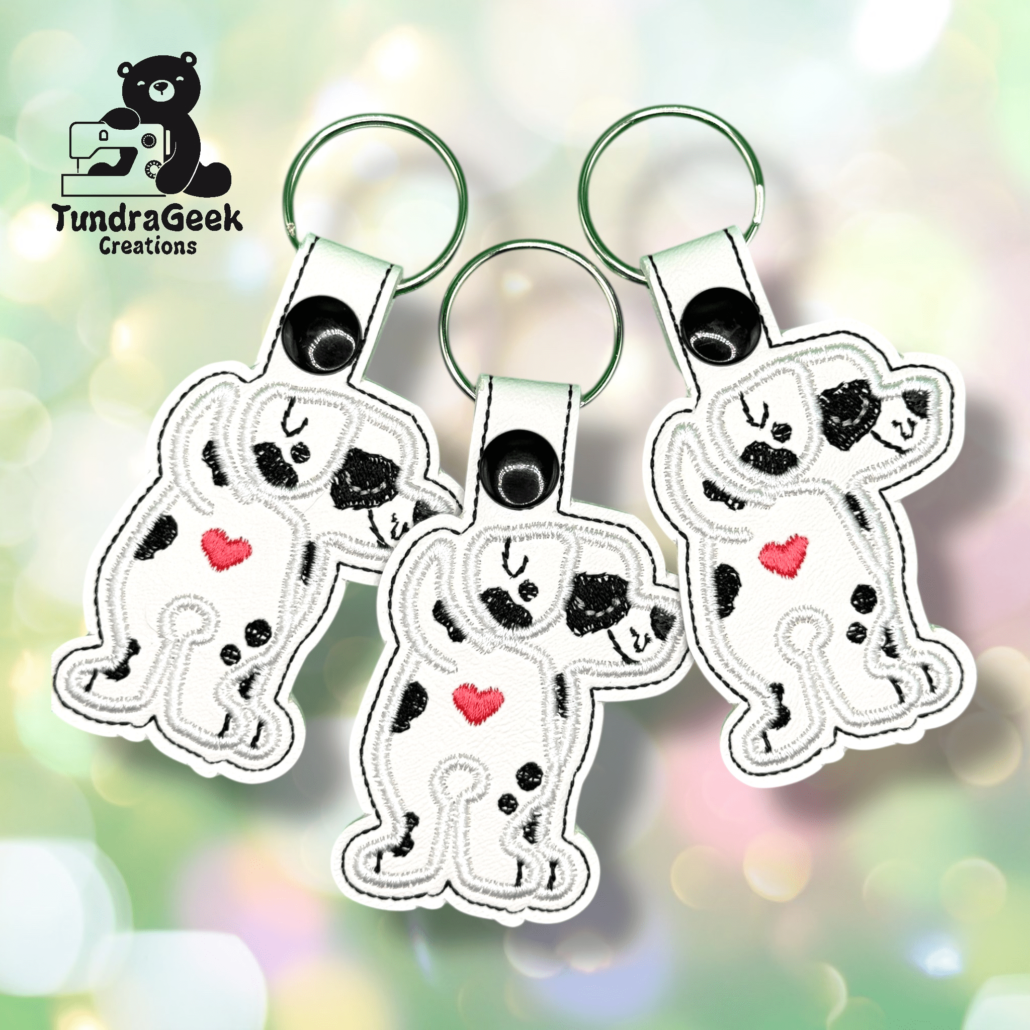 Three embroidered keychains featuring a cartoon Dalmatian with a red heart on its chest, each in a dabbing pose, are displayed on a pastel background with the TundraGeek Creations logo.