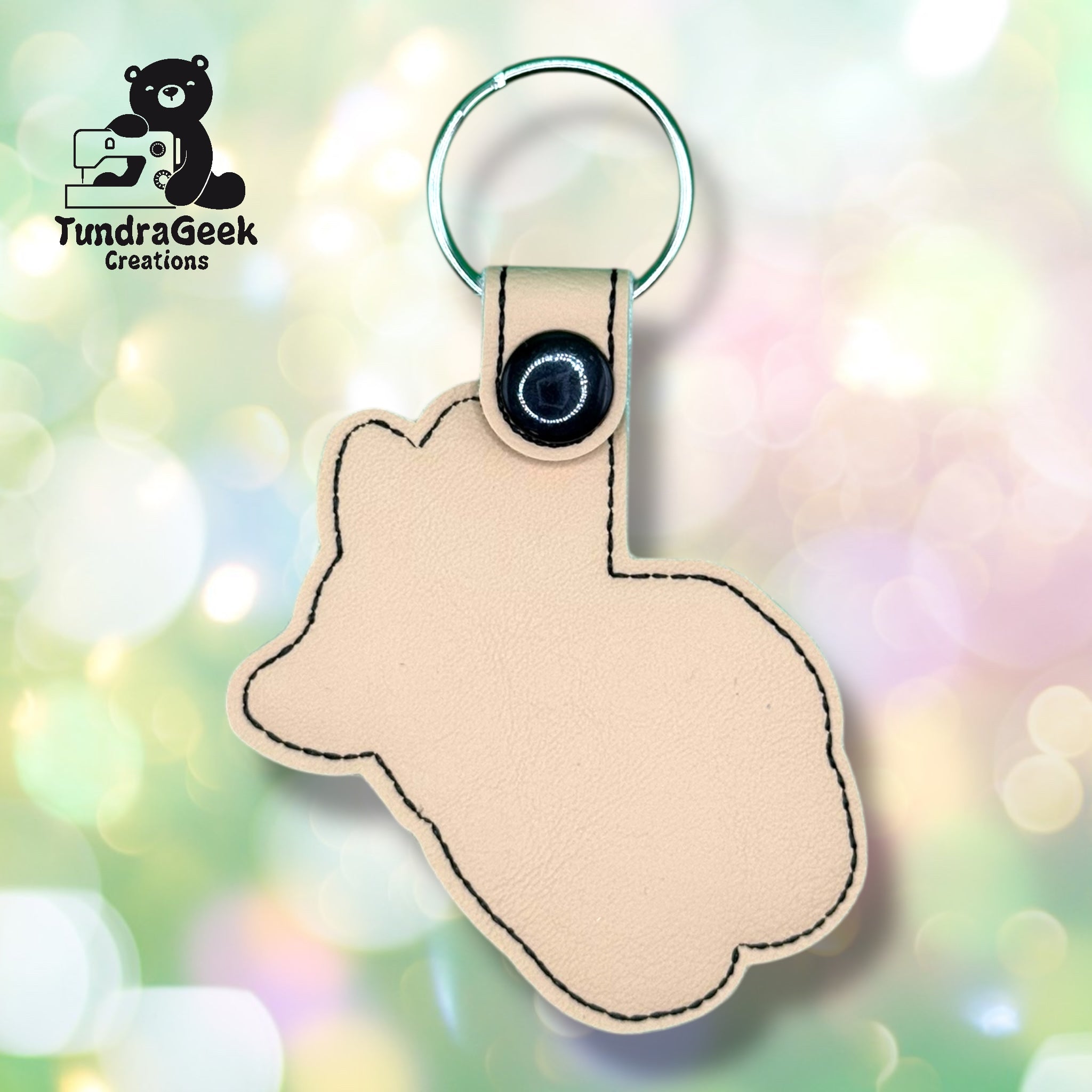 A beige keychain shaped like a rabbit with a metal ring and a snap button, displayed on a soft, colorful, blurred background. TundraGeek Creations logo is visible in the corner.