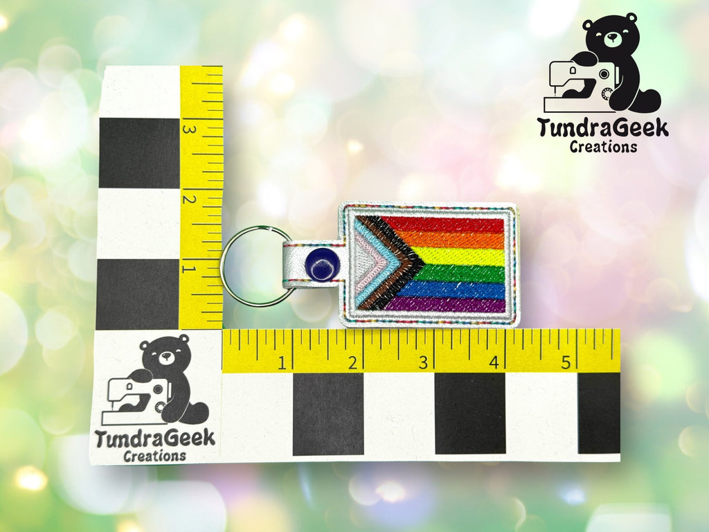 A rectangular rainbow Pride keychain with a blue snap is placed on a measuring grid showing TundraGeek Creations branding. The keychain measures about 2 inches wide and 1.5 inches tall.