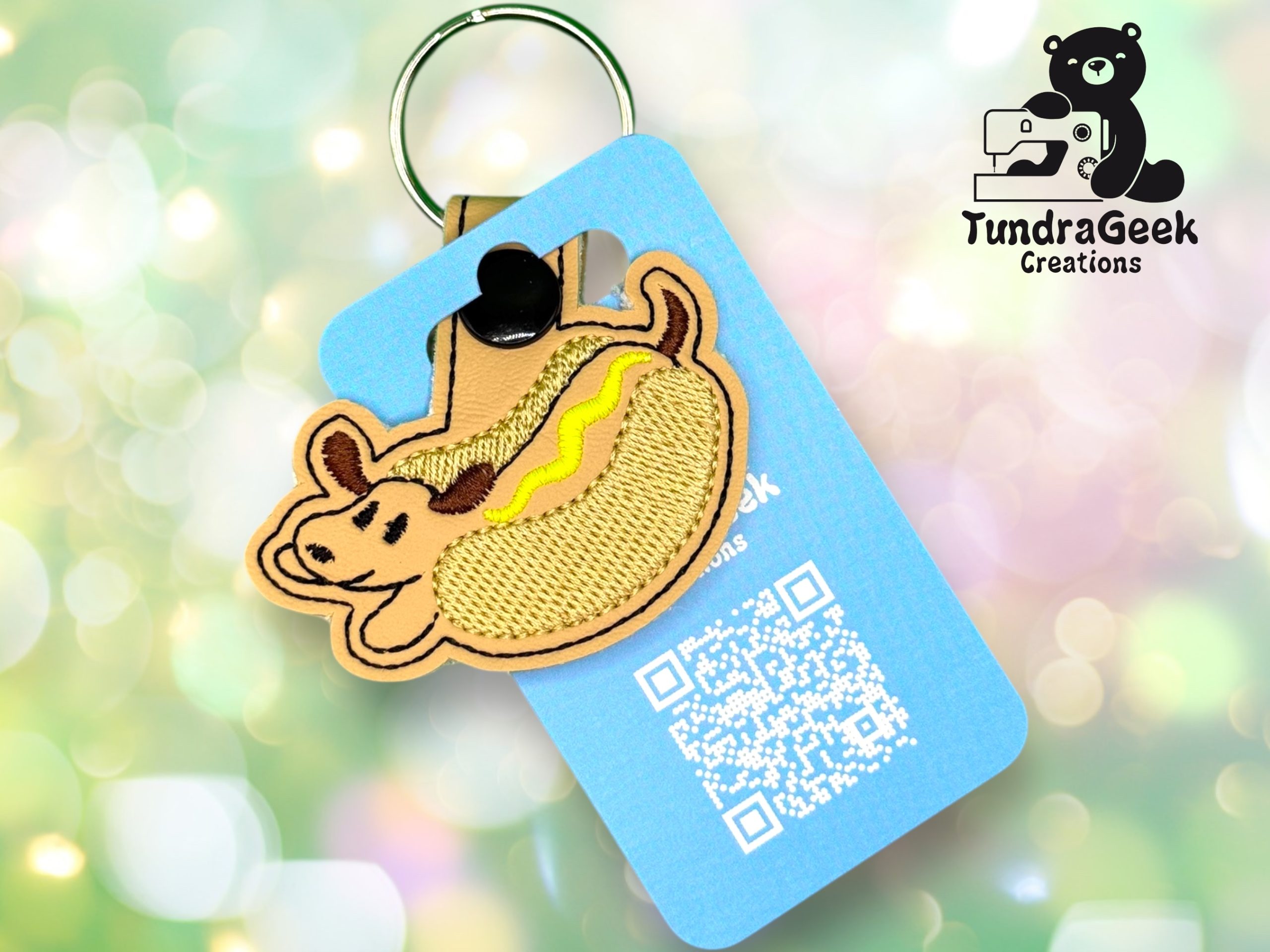 A keychain with a felt dog in a hot dog bun design attached to a blue card with a QR code and the TundraGeek Creations logo in the background.