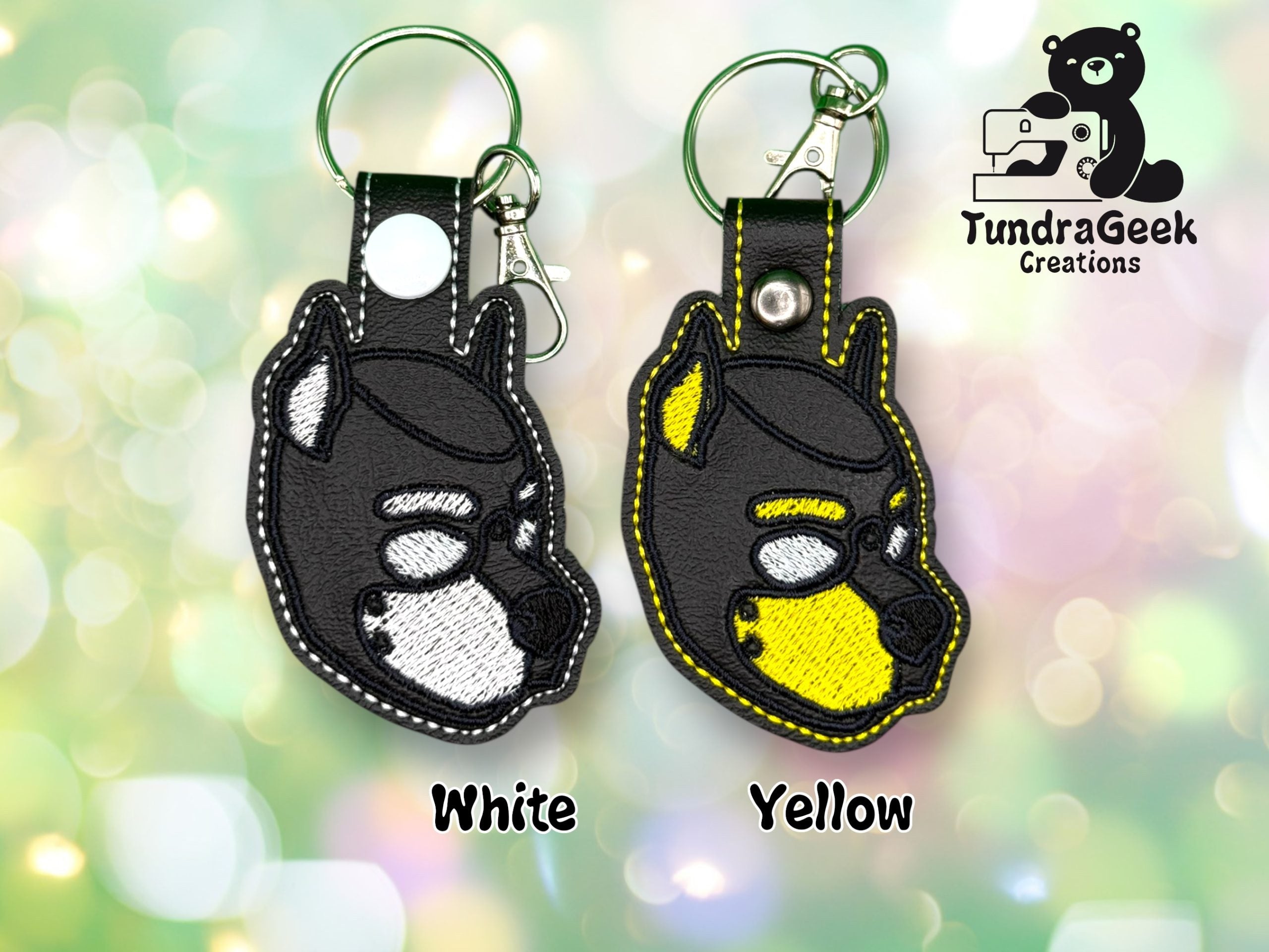 Two dog-shaped keychains, one with a white snout and one with a yellow snout, displayed on a blurred green background with the TundraGeek Creations logo in the top right corner.