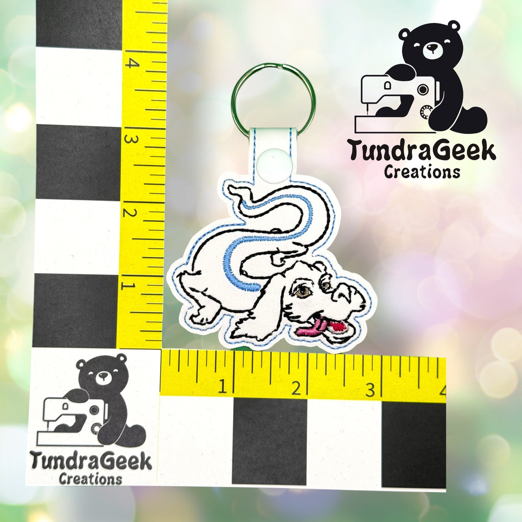 A keychain featuring a white flying dragon with blue accents, placed on a measurement grid with TundraGeek Creations logos in the background.