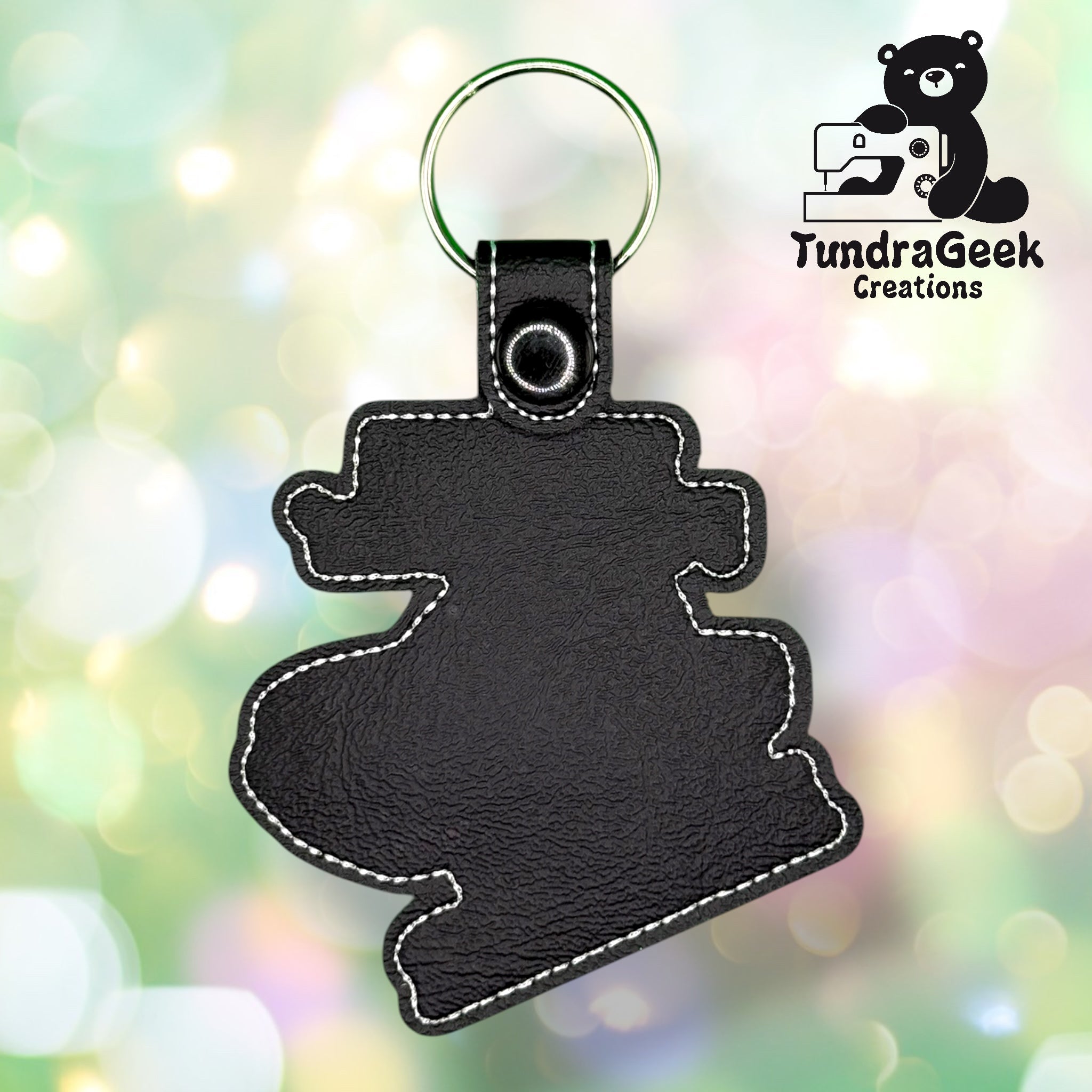 Black leather keychain in the shape of a stylized cartoon hippo, displayed against a blurred pastel background with the TundraGeek Creations logo in the top right corner.