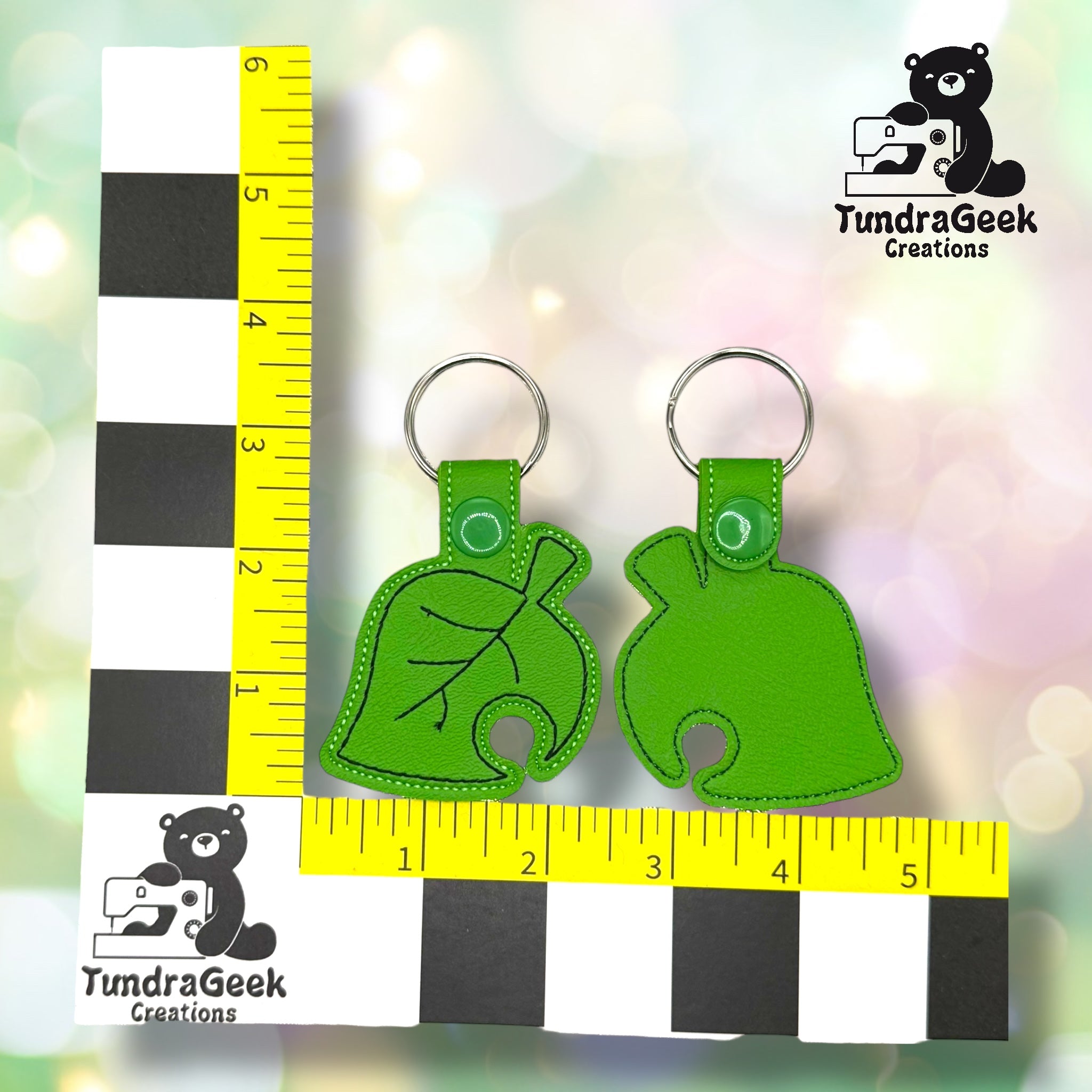 Two green keychains shaped like a leaf and a stylized animal crossing leaf, displayed on a yellow and black measurement grid with TundraGeek Creations logos.