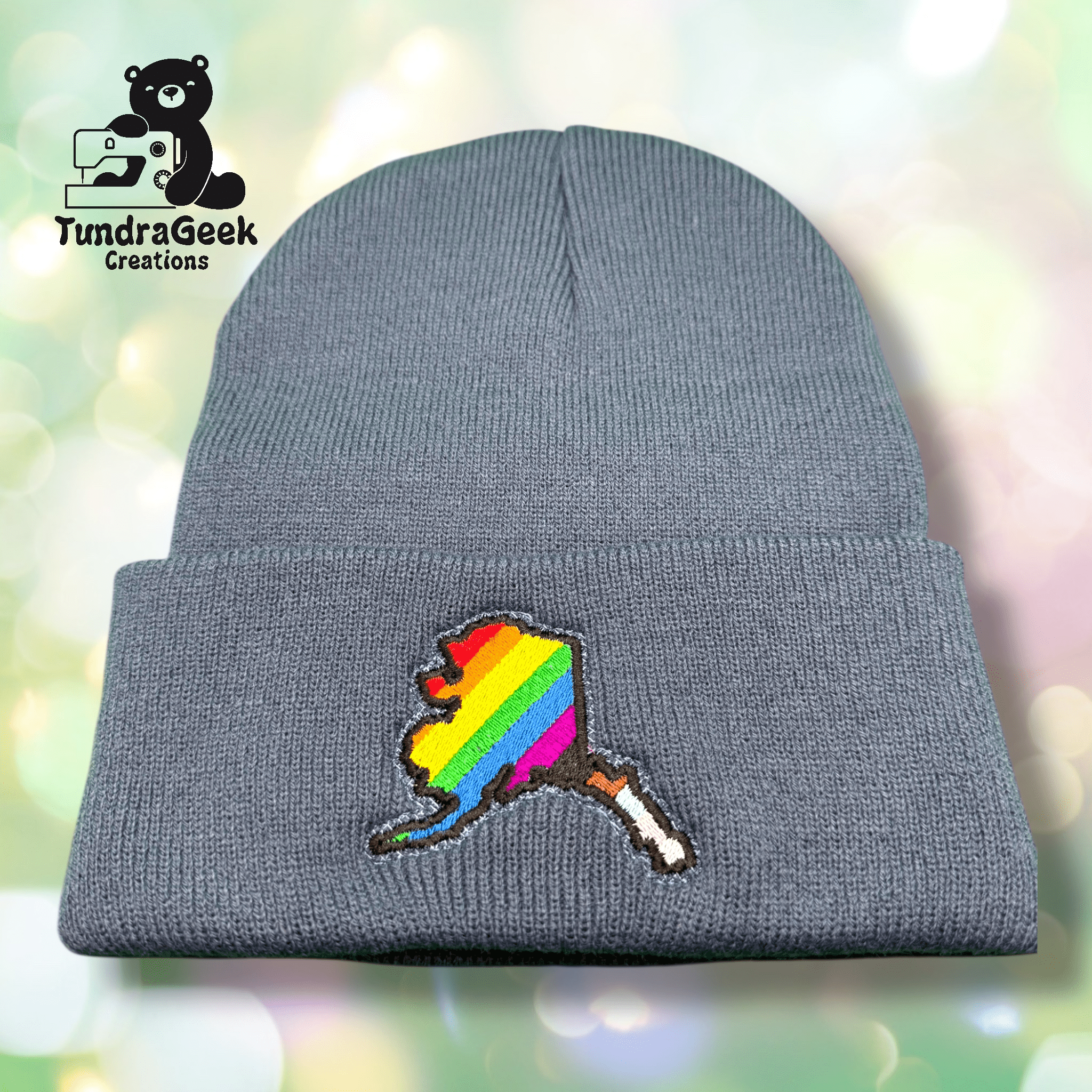 Gray knit beanie featuring a stitched design of a leaf with rainbow stripes; TundraGeek Creations logo in top left corner.