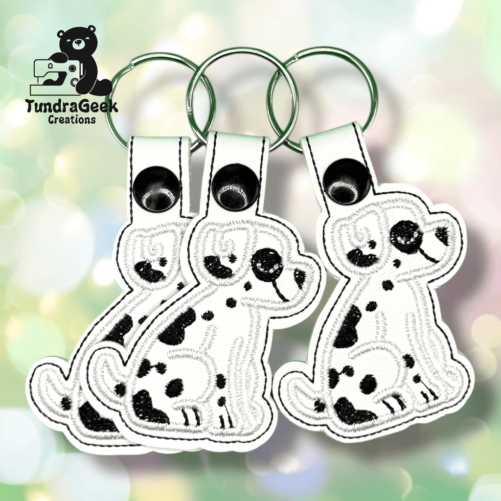 Three identical spotted dog keychains made of white material with black stitching are displayed side by side on a pastel bokeh background. “TundraGeek Creations” logo appears in the top left corner.