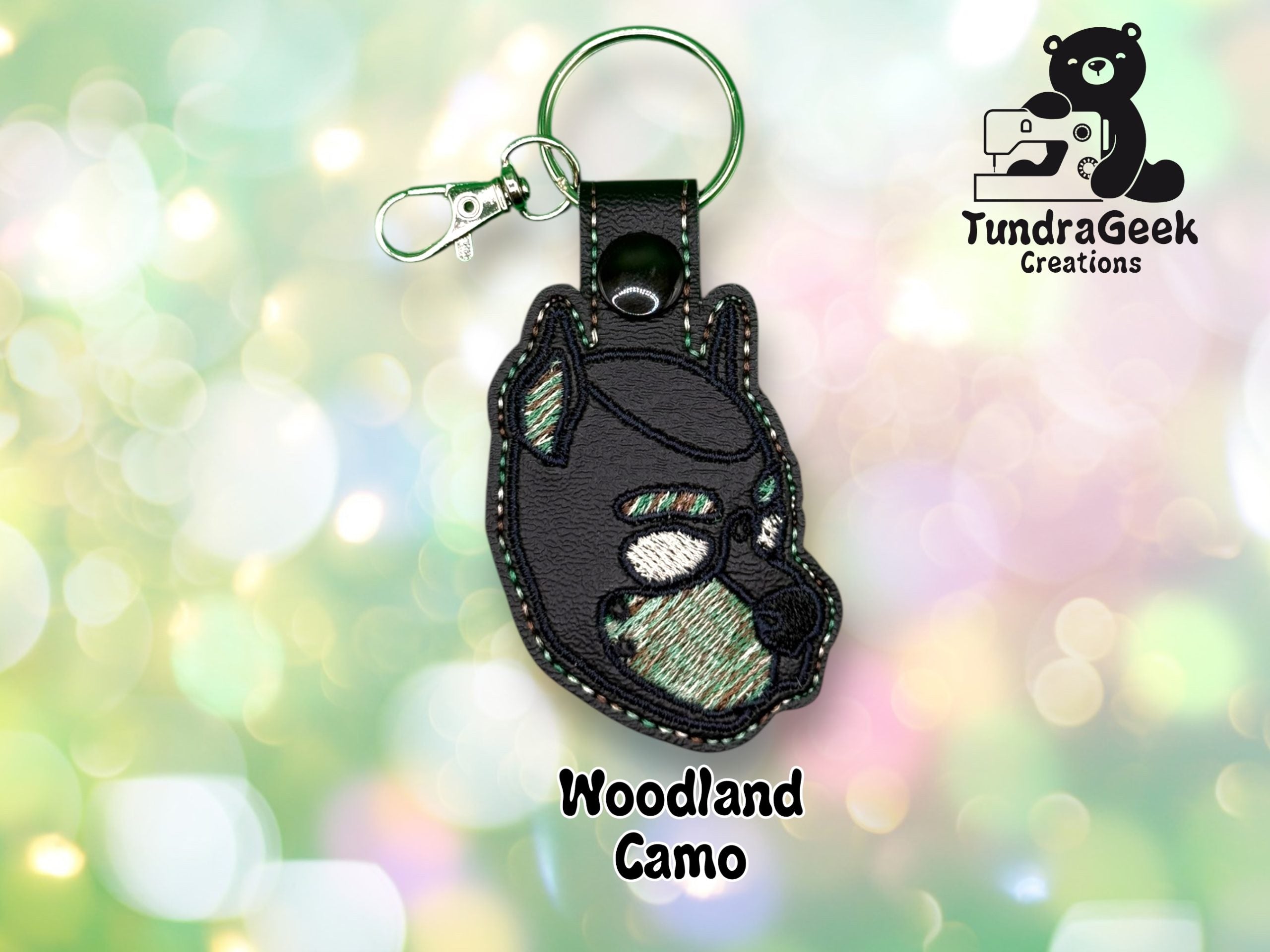 Keychain featuring an embroidered bear face in woodland camo colors, displayed on a blurred green background with the TundraGeek Creations logo.