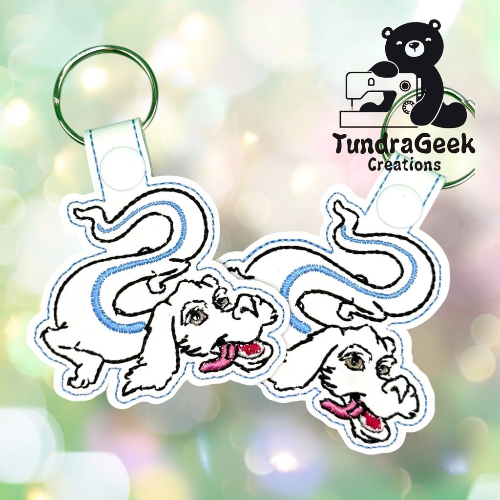 Two embroidered white dragon keychains with blue accents on split rings, displayed in front of a bokeh background, with the TundraGeek Creations logo in the top right corner.
