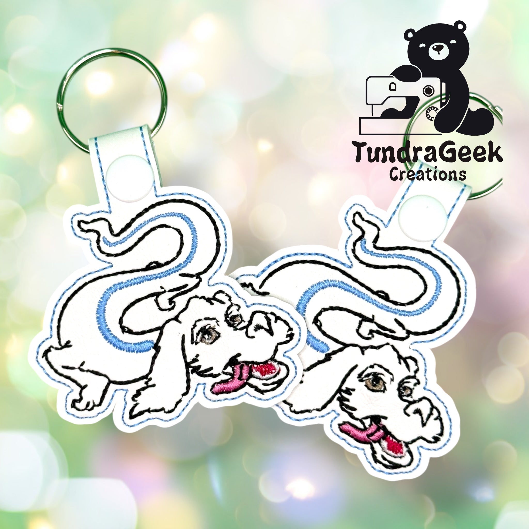 Two embroidered white dragon keychains with blue accents on split rings, displayed in front of a bokeh background, with the TundraGeek Creations logo in the top right corner.