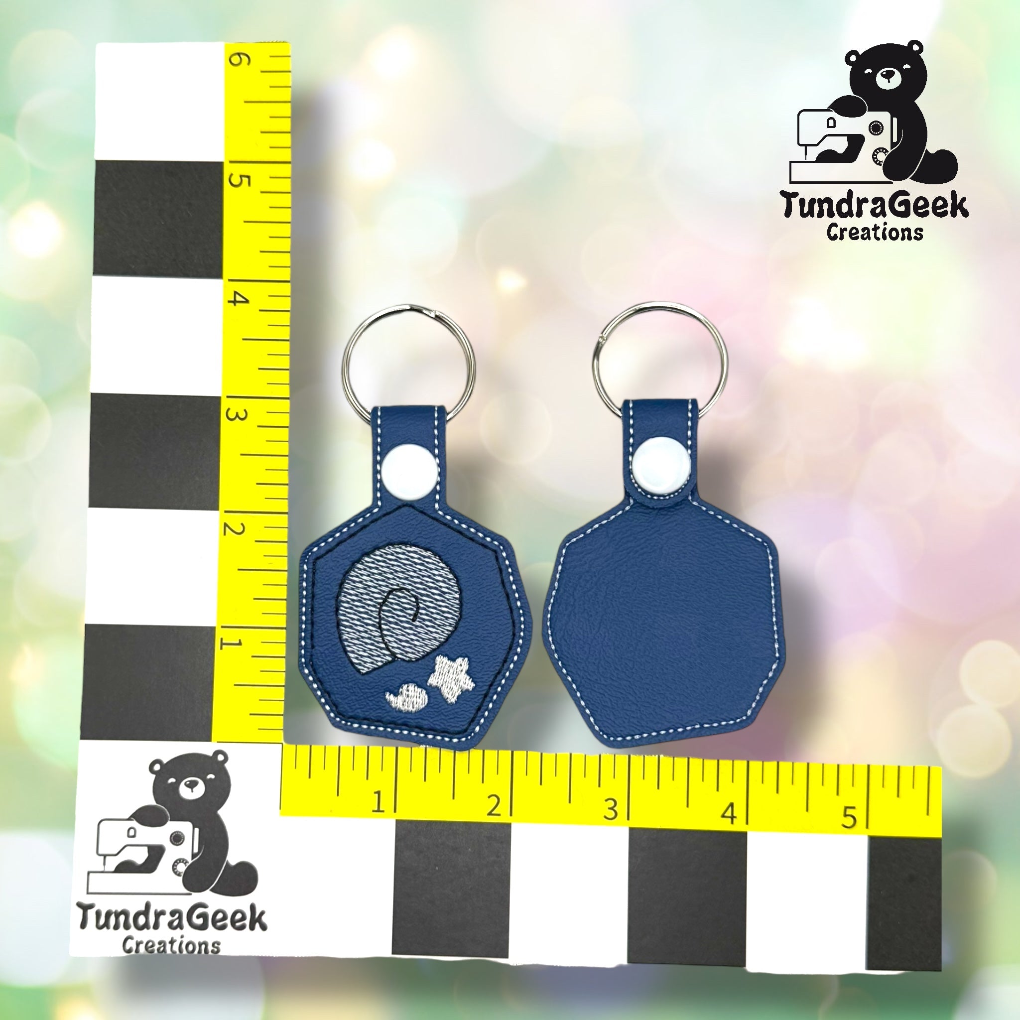 Two blue leather keychains, one with a seashell and starfish design, displayed on a measurement grid with a TundraGeek Creations logo in the background.