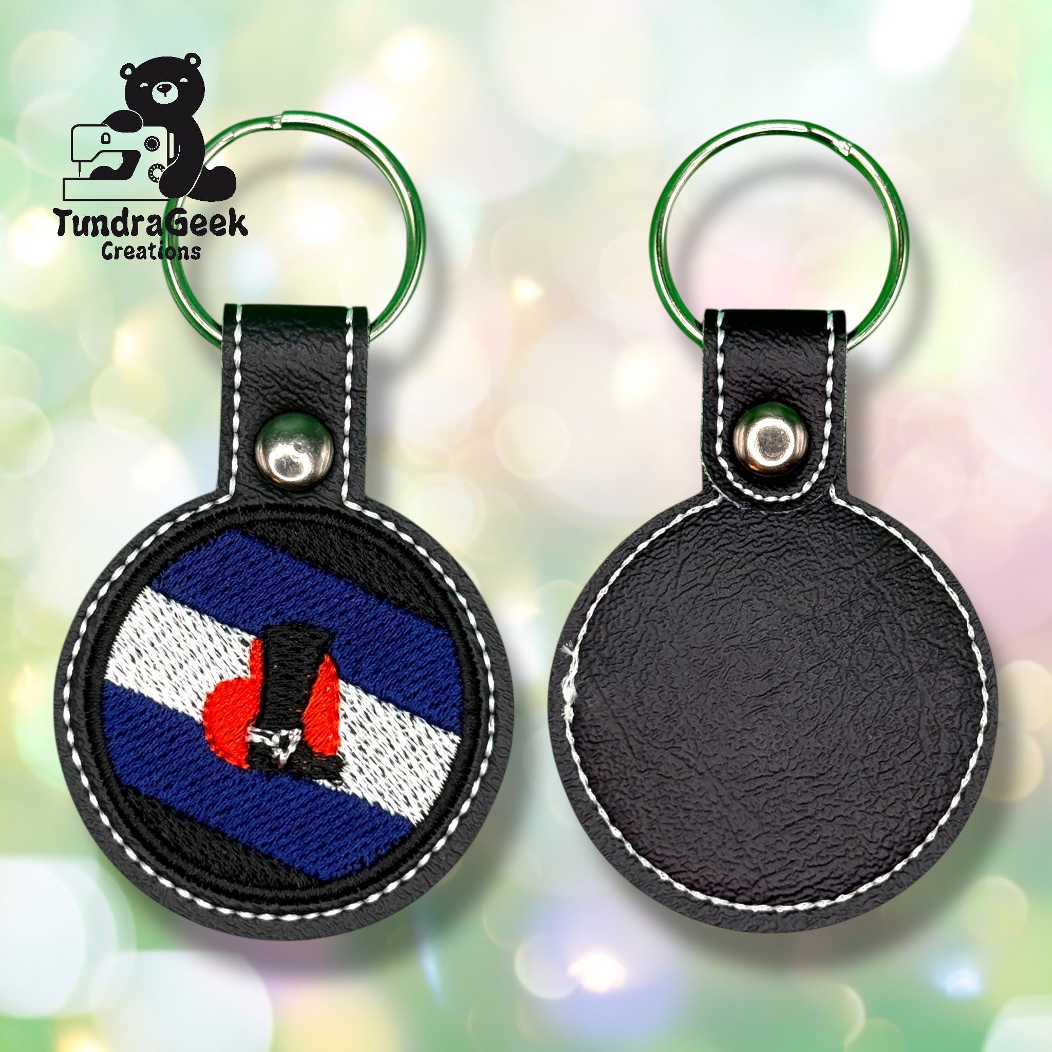 Two round leather keychains, one with a blue, black, white, and red embroidered flag design and one plain, displayed on a blurred background with the Tundra Geek Creations logo.