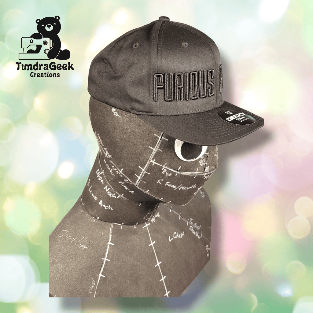 A gray hat with FURIOUS embroidered on it sits on a brown mannequin head marked with sewing pattern labels. The TundraGeek Creations logo appears in the top left corner.