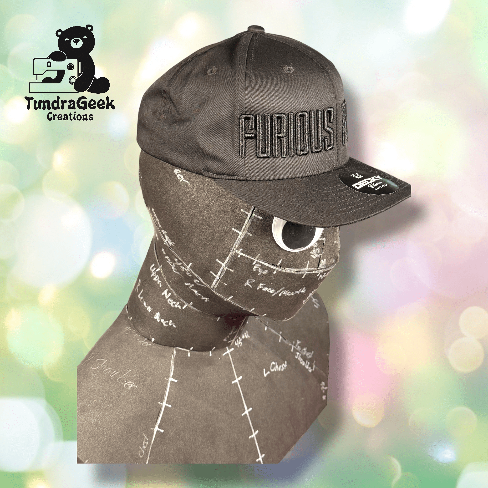 A gray hat with FURIOUS embroidered on it sits on a brown mannequin head marked with sewing pattern labels. The TundraGeek Creations logo appears in the top left corner.