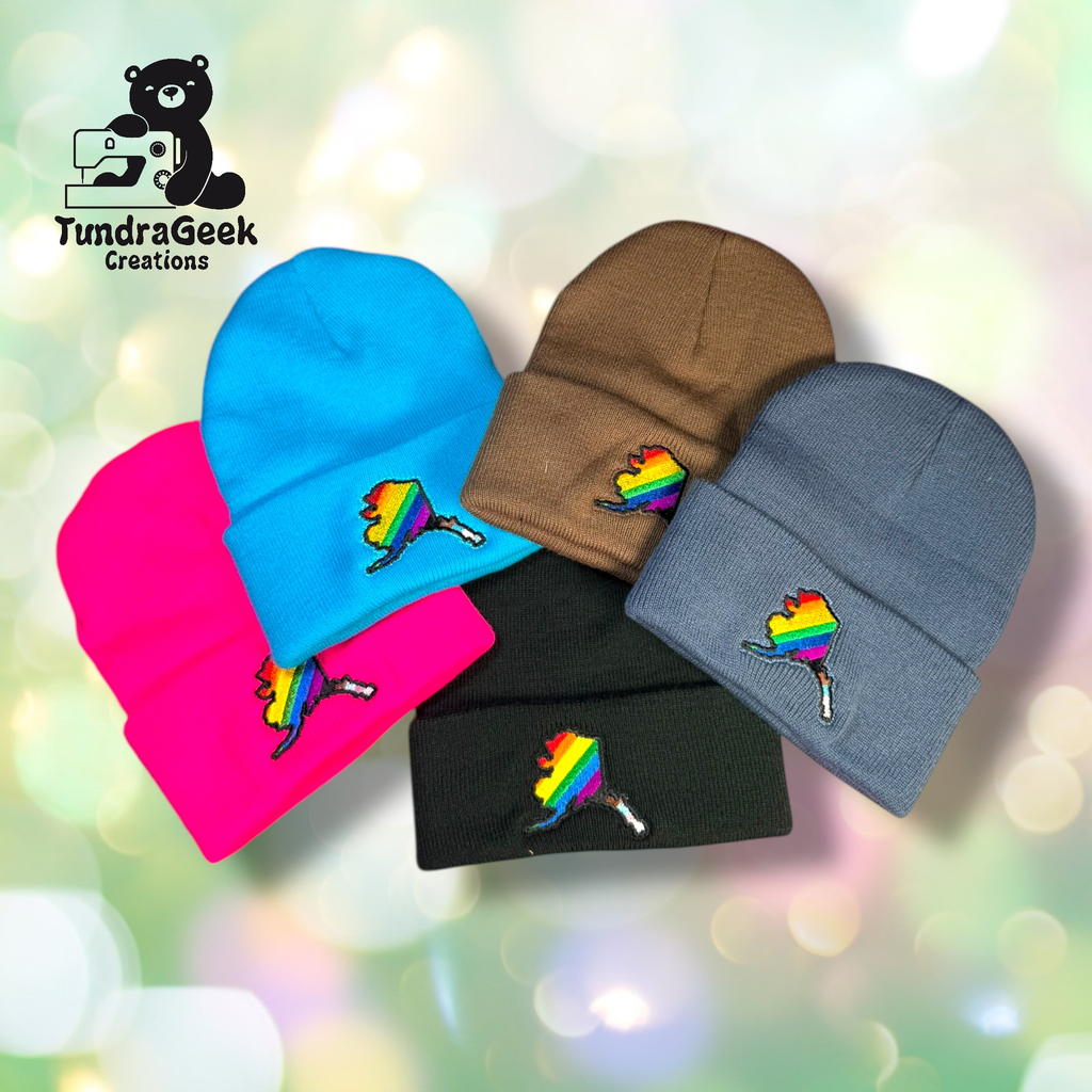 Five knit beanies in blue, brown, gray, pink, and black, each embroidered with an Alaska state outline in rainbow colors, displayed on a soft-focus background. TundraGeek Creations logo in corner.