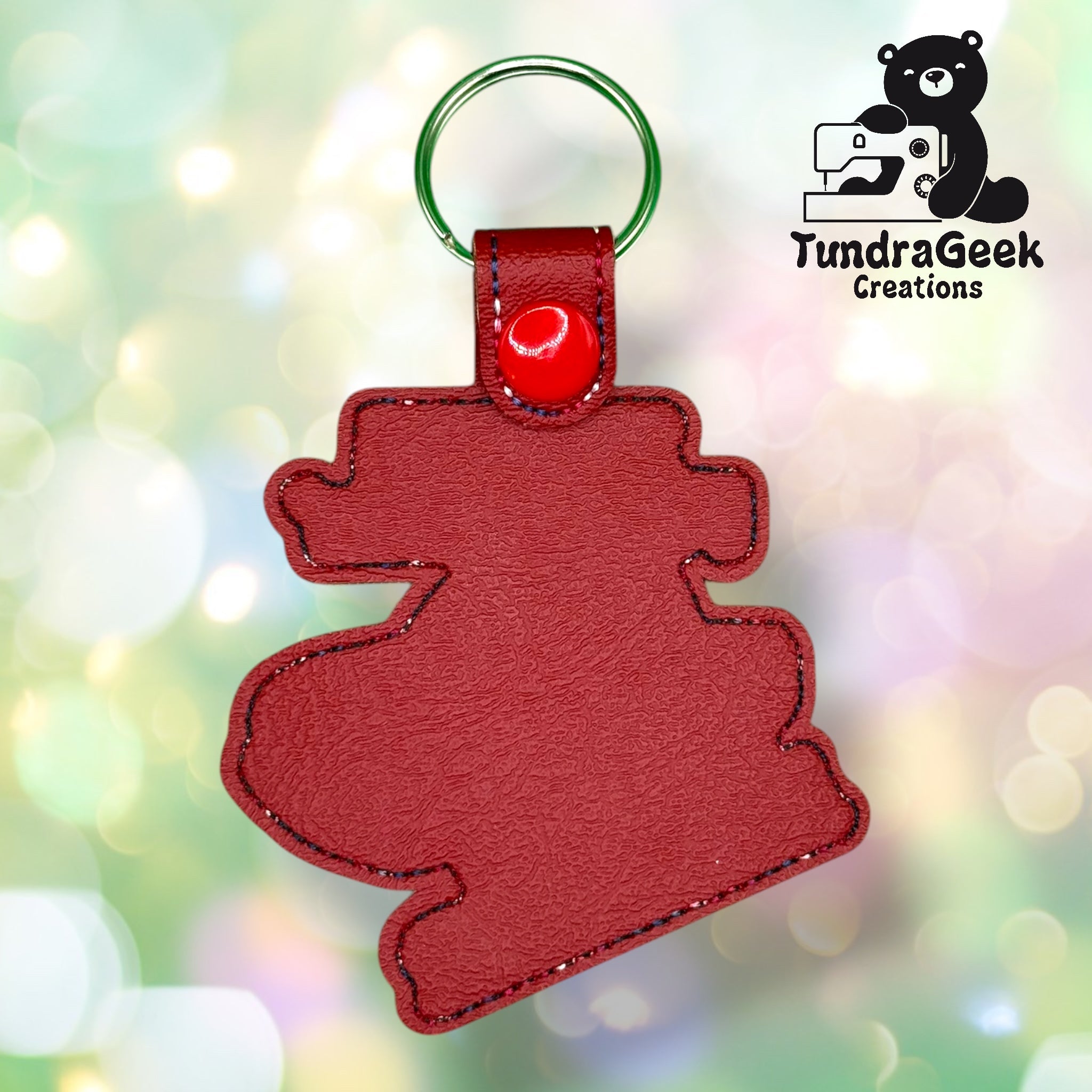 Red leather keychain in the shape of a cartoon-like figure with a green keyring and red snap, displayed against a blurred, colorful background. TundraGeek Creations logo in the corner.