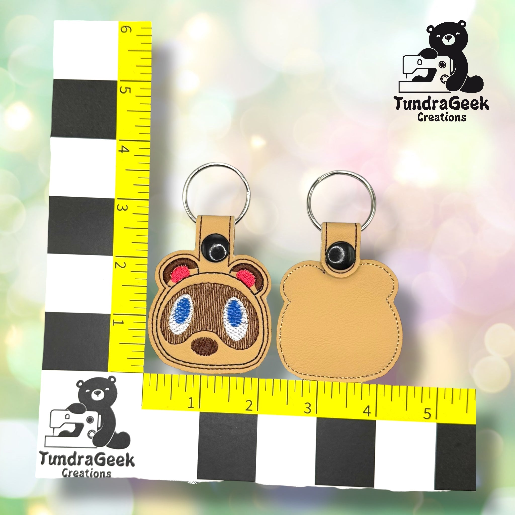Two brown keychains, one with an embroidered animal face and one plain, are displayed on a yellow ruler grid next to the TundraGeek Creations logo.