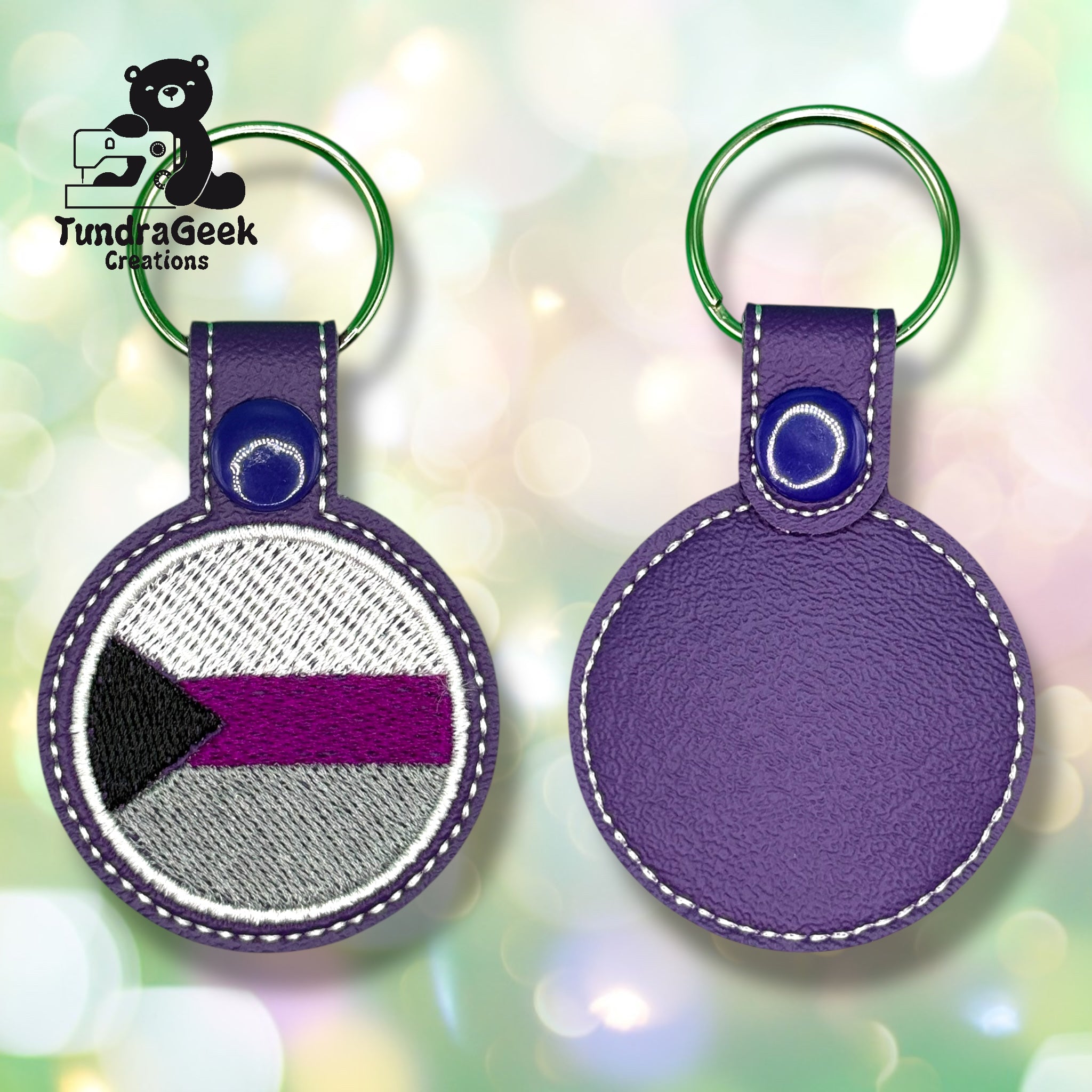 Two round purple keychains, one showing an embroidered pride flag with black, gray, white, and purple stripes, the other showing the plain back.