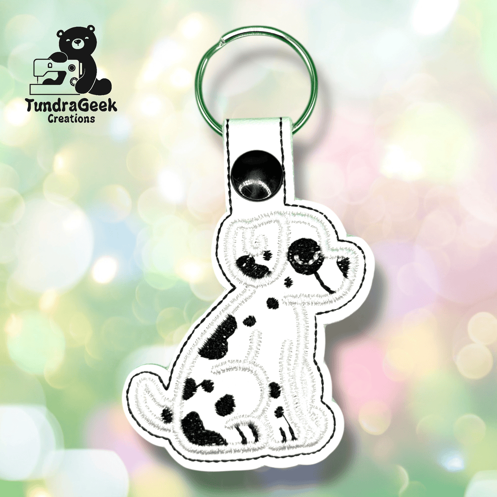 A white keychain shaped like a cartoon Dalmatian dog with black spots is displayed on a blurred pastel background. The TundraGeek Creations logo appears in the top left corner.