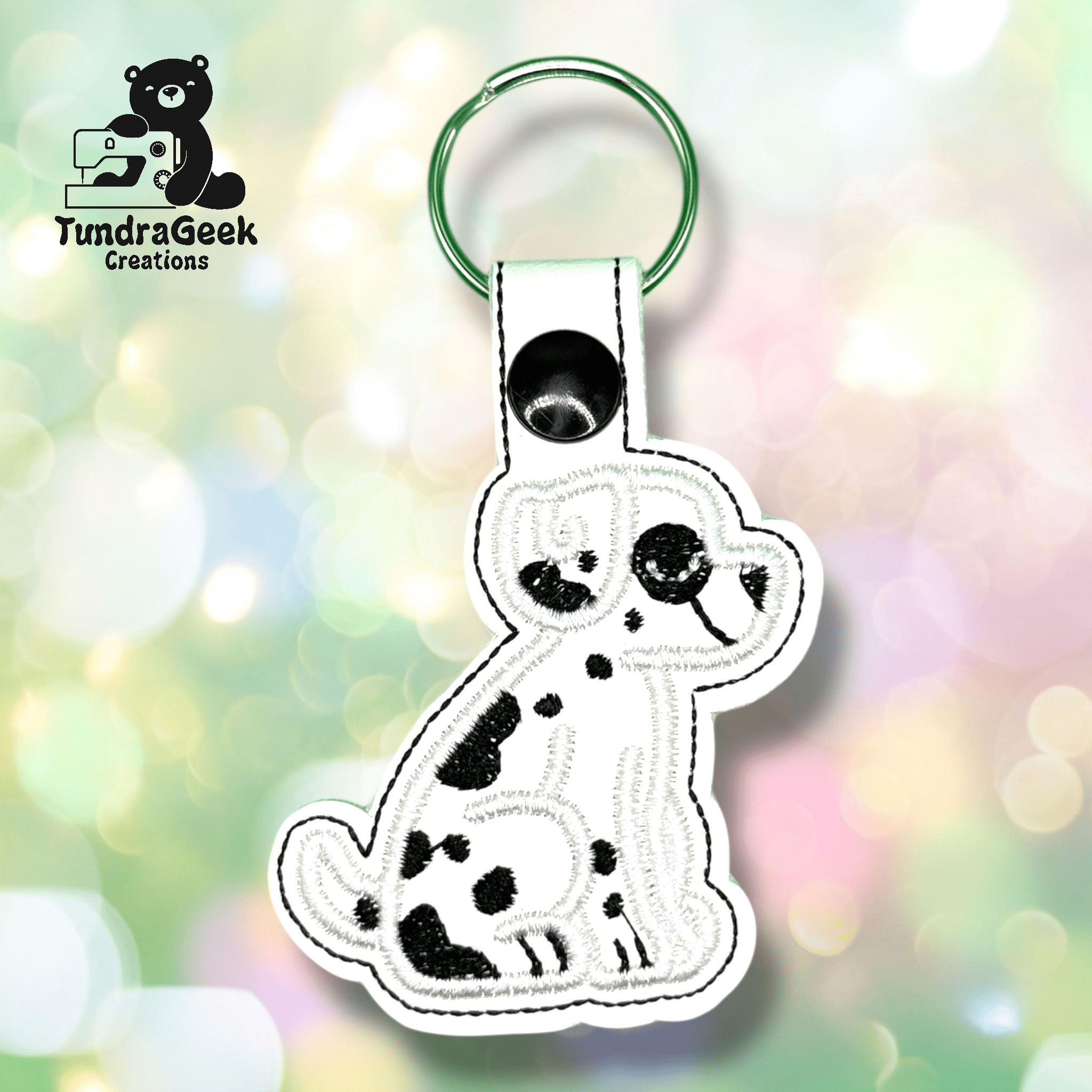 A white keychain shaped like a cartoon Dalmatian dog with black spots is displayed on a blurred pastel background. The TundraGeek Creations logo appears in the top left corner.