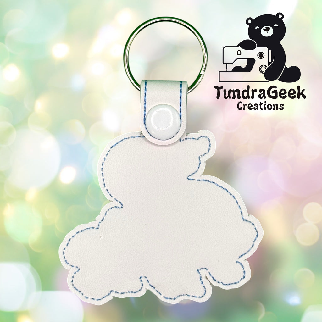 White, bunny-shaped faux leather keychain with blue stitching, shown against a blurred pastel background. TundraGeek Creations logo is in the top right corner.