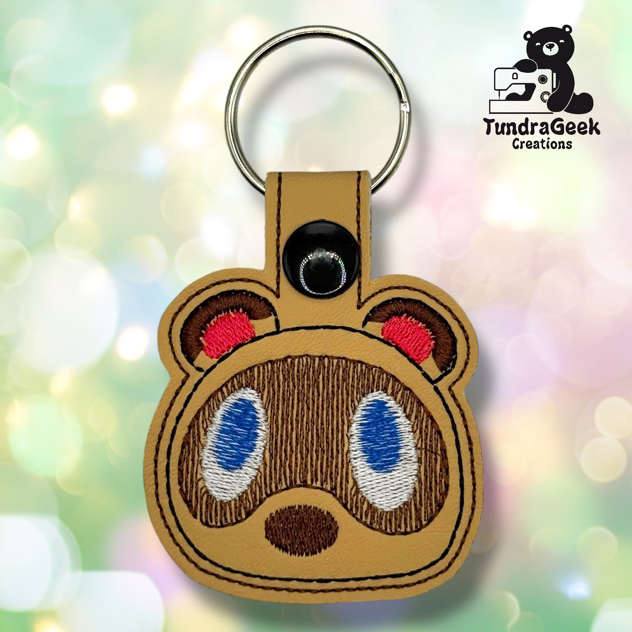 Brown bear face keychain with embroidered blue, white, and brown details, a metal keyring, and the TundraGeek Creations logo on a blurred pastel background.