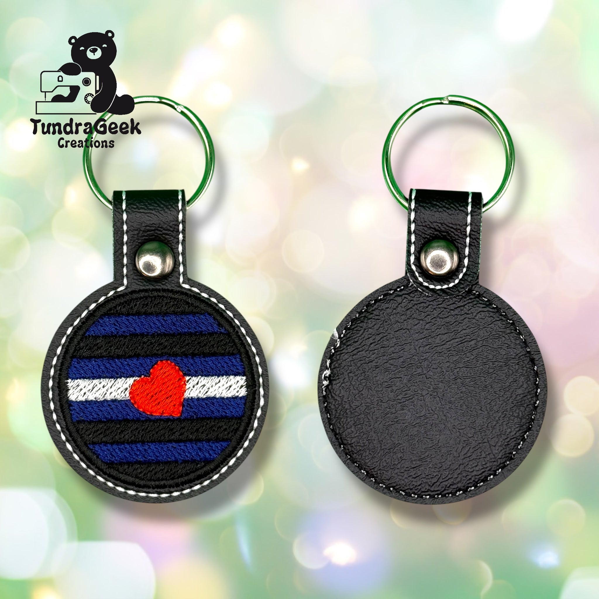 Two round keychains with green keyrings; one shows a leather pride flag with a red heart, the other is plain black leather. TundraGeek Creations logo is in the top left corner.