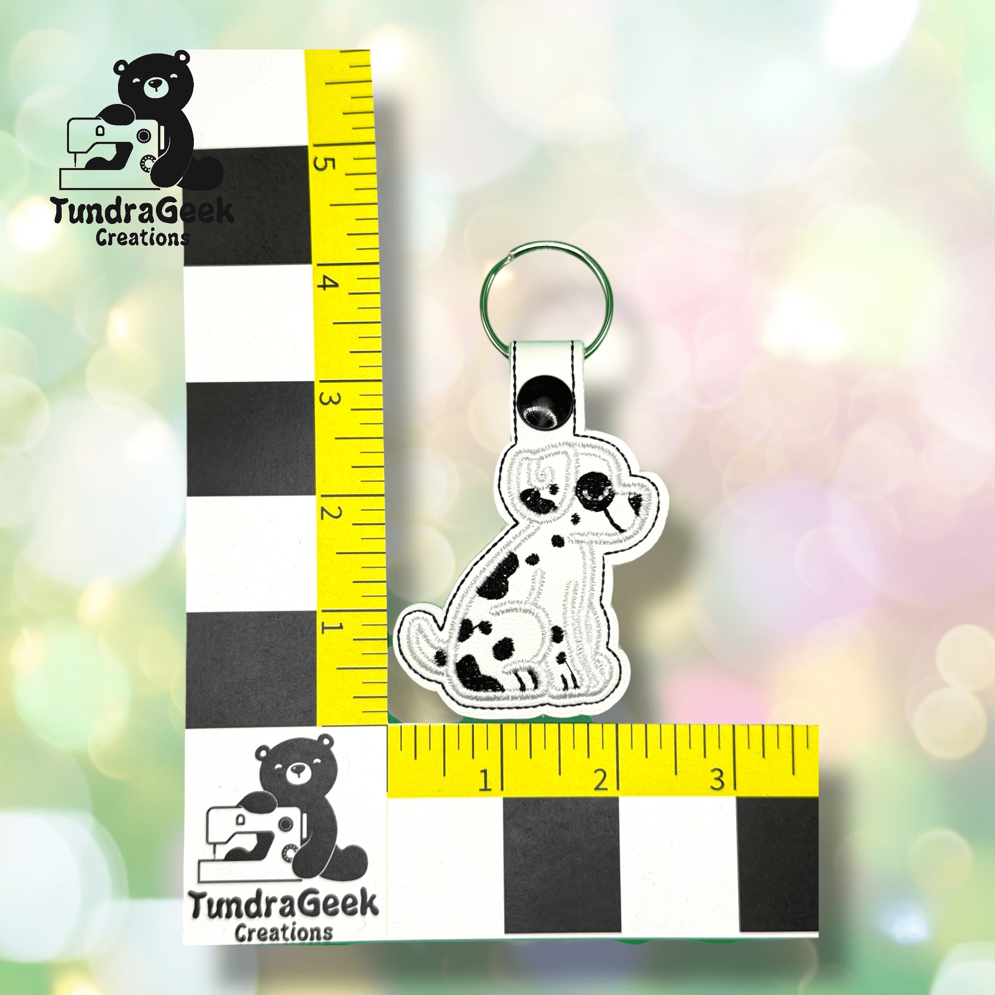 A white and black dog-shaped keychain is displayed on a measurement grid, showing its about 3 inches tall. TundraGeek Creations logo appears twice on the grid.