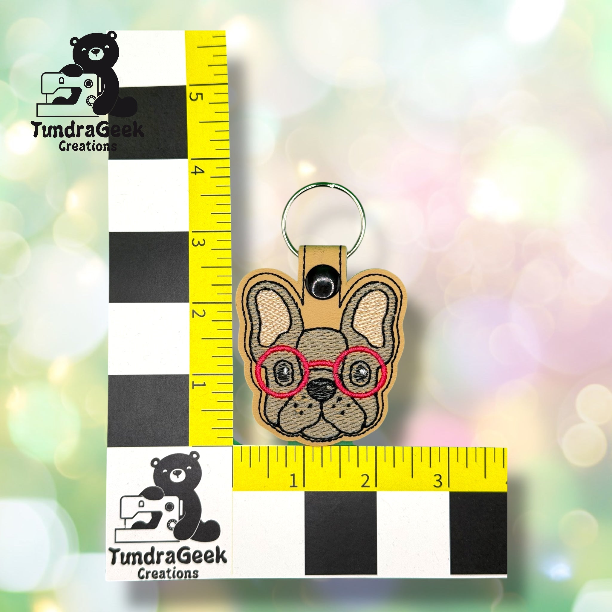 A keychain featuring a French bulldog with red glasses, placed between two measuring rulers on a blurred background with TundraGeek Creations logos.