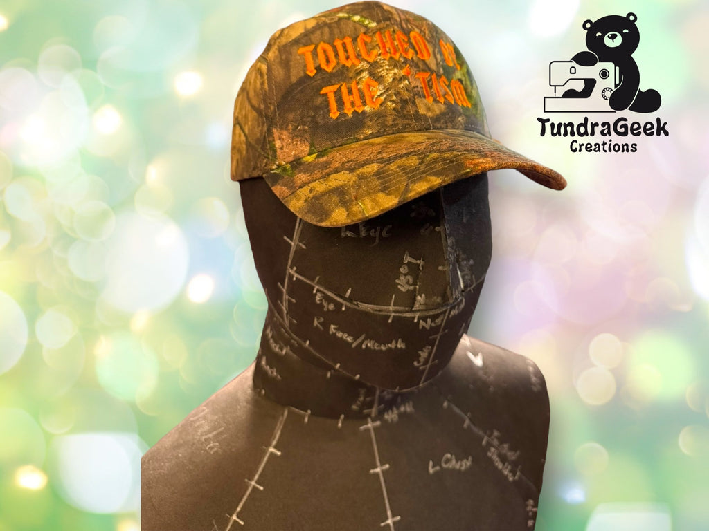 A mannequin head displays a camouflage hat with orange text. The background is green with bokeh, and the TundraGeek Creations logo is in the upper right corner.