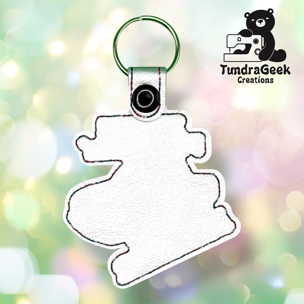 A blank white keychain with a stitched outline, shaped like a cartoon character, on a blurred bokeh background. TundraGeek Creations logo is in the top right corner.