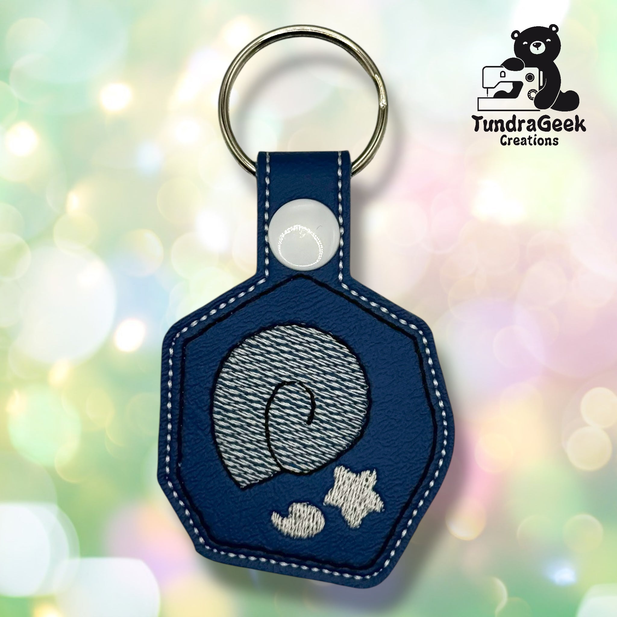 A blue hexagonal keychain with embroidered gray ammonite fossil design, attached to a metal keyring. A black bear sewing logo and TundraGeek Creations text appear in the top right.