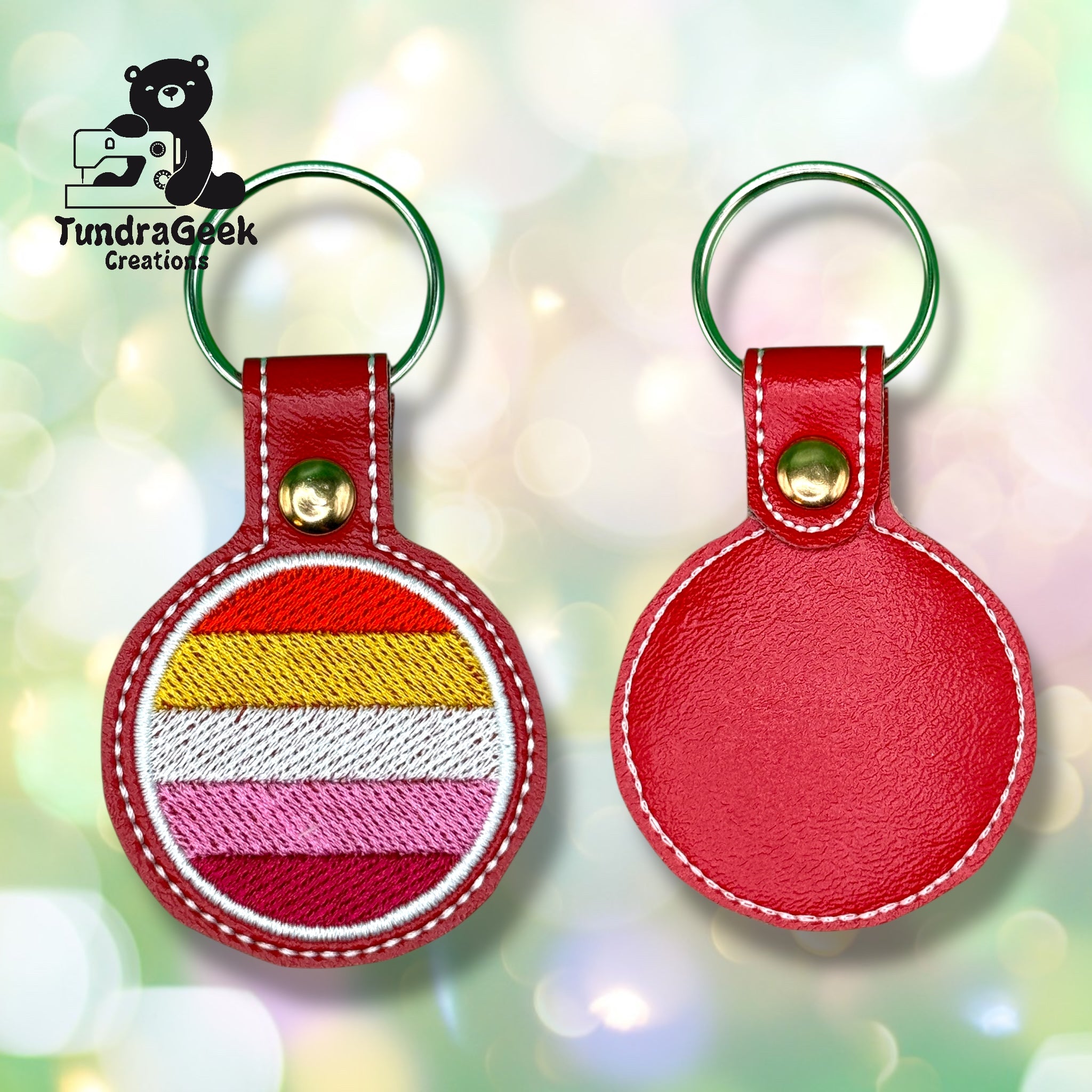 Two red leather keychains; one shows horizontal stripes in yellow, white, orange, pink, and red, while the other shows the plain back. TundraGeek Creations logo in corner.