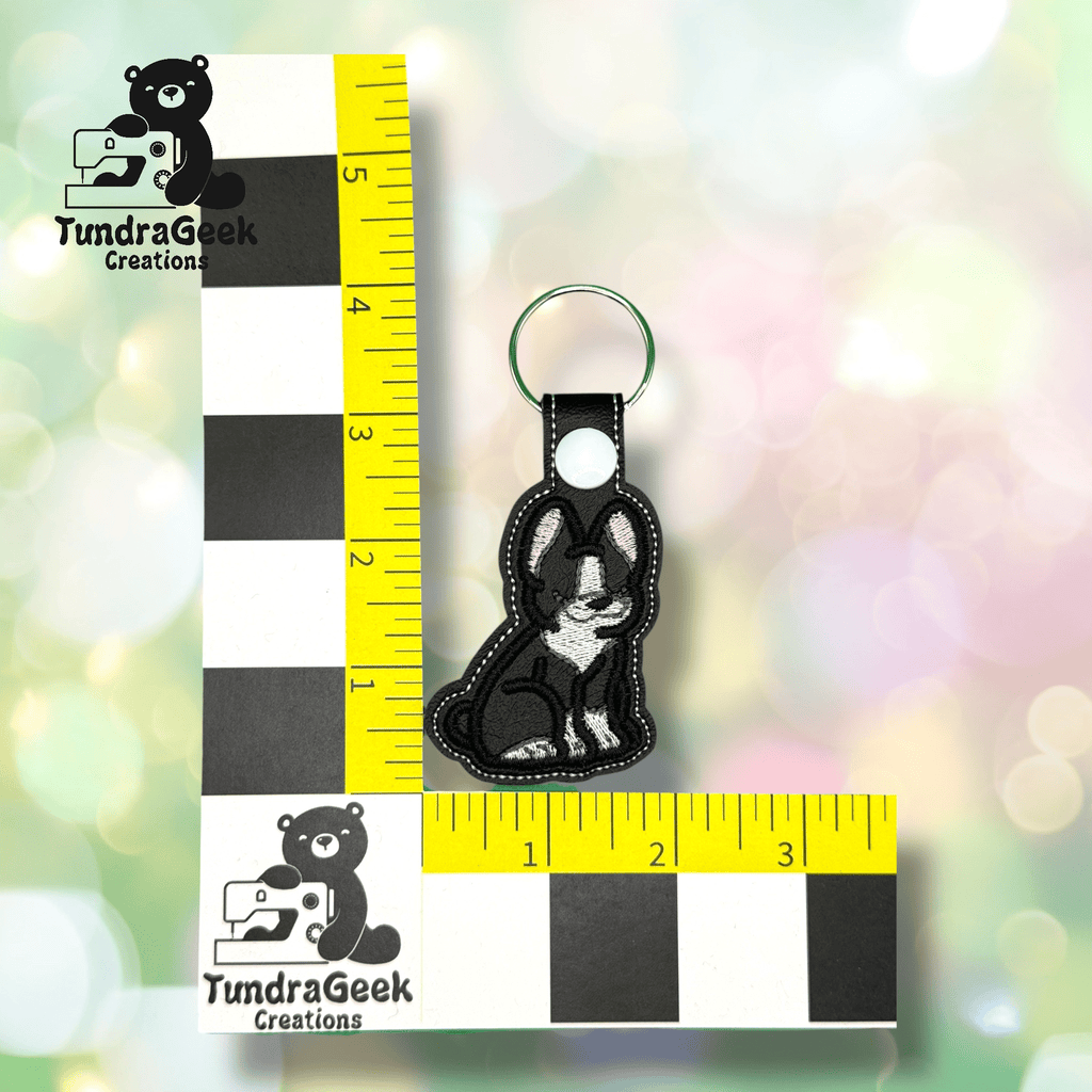 Keychain featuring an embroidered black and white dog placed next to a ruler for scale, measuring approximately 3 inches tall. TundraGeek Creations logo visible on the rulers.