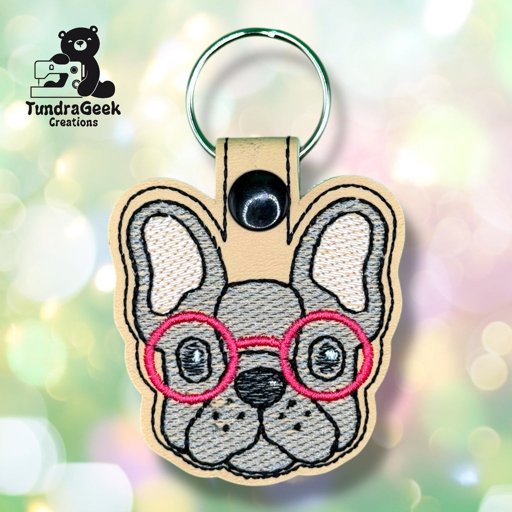 A keychain shaped like a French bulldogs face, embroidered with red glasses, and a TundraGeek Creations logo in the top left corner.