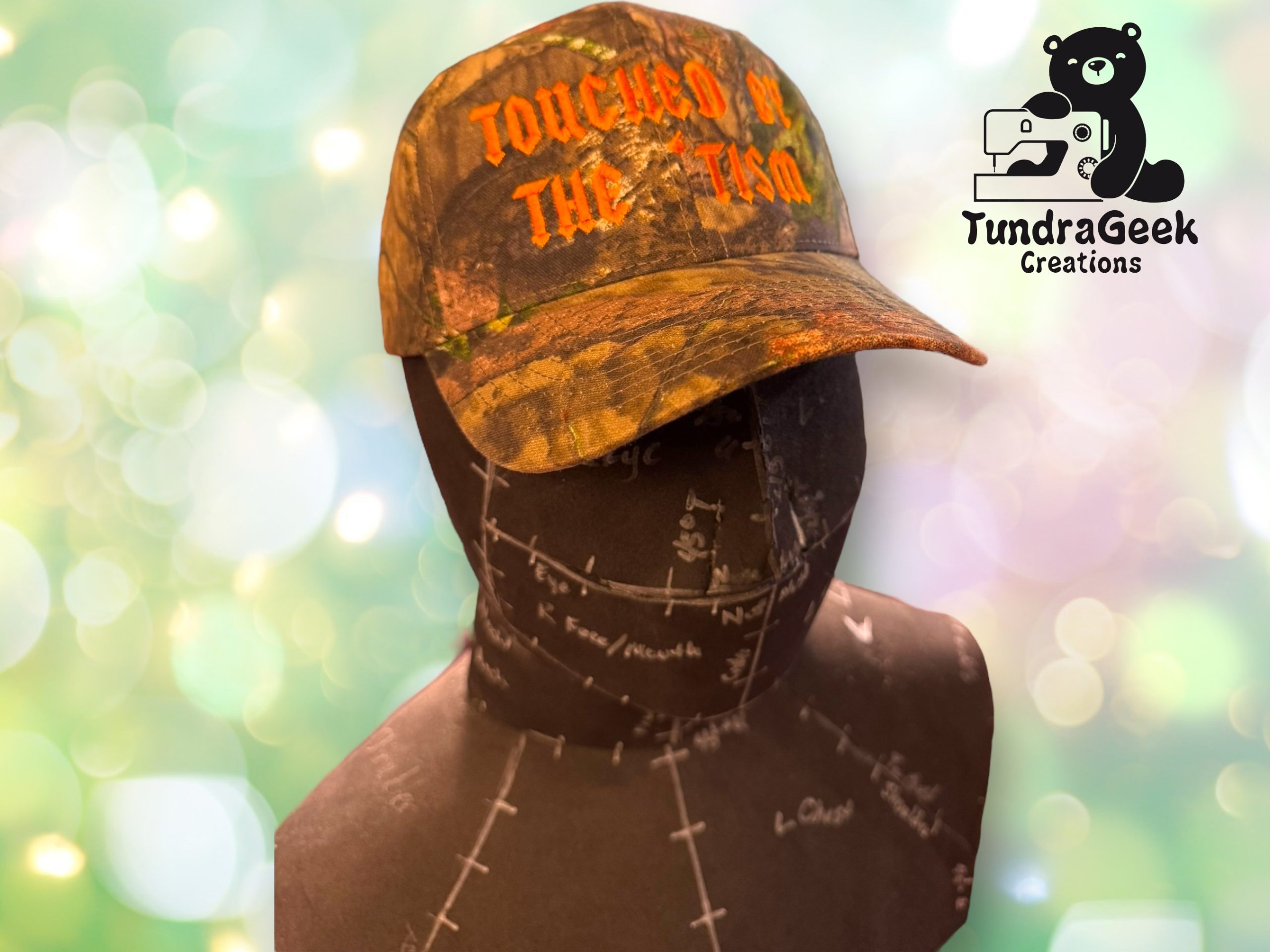 A mannequin displays a camo hat with TOUCHED BY THE TUNDRA in orange letters and a black neck gaiter printed with white maps; TundraGeek Creations logo is in the corner.