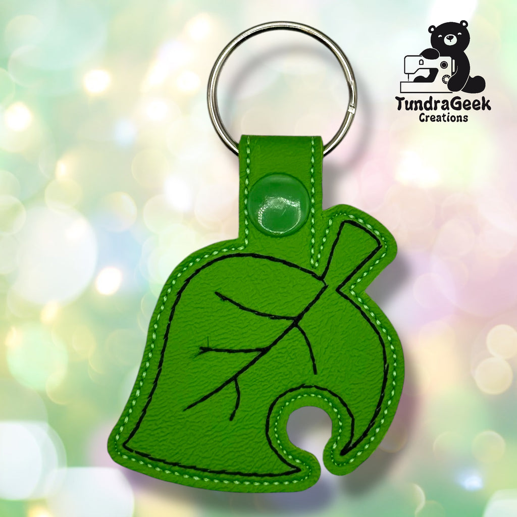 A green leaf-shaped keychain with black stitching, a snap button, and a keyring, displayed on a blurred light background with the TundraGeek Creations logo in the corner.