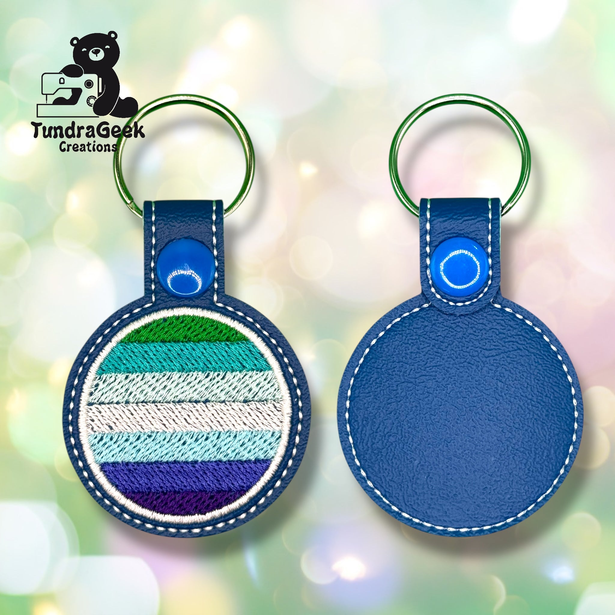 Two round blue keychains; one side is plain, the other features striped embroidery in green, white, gray, and purple. The TundraGeek Creations logo is visible in the background.