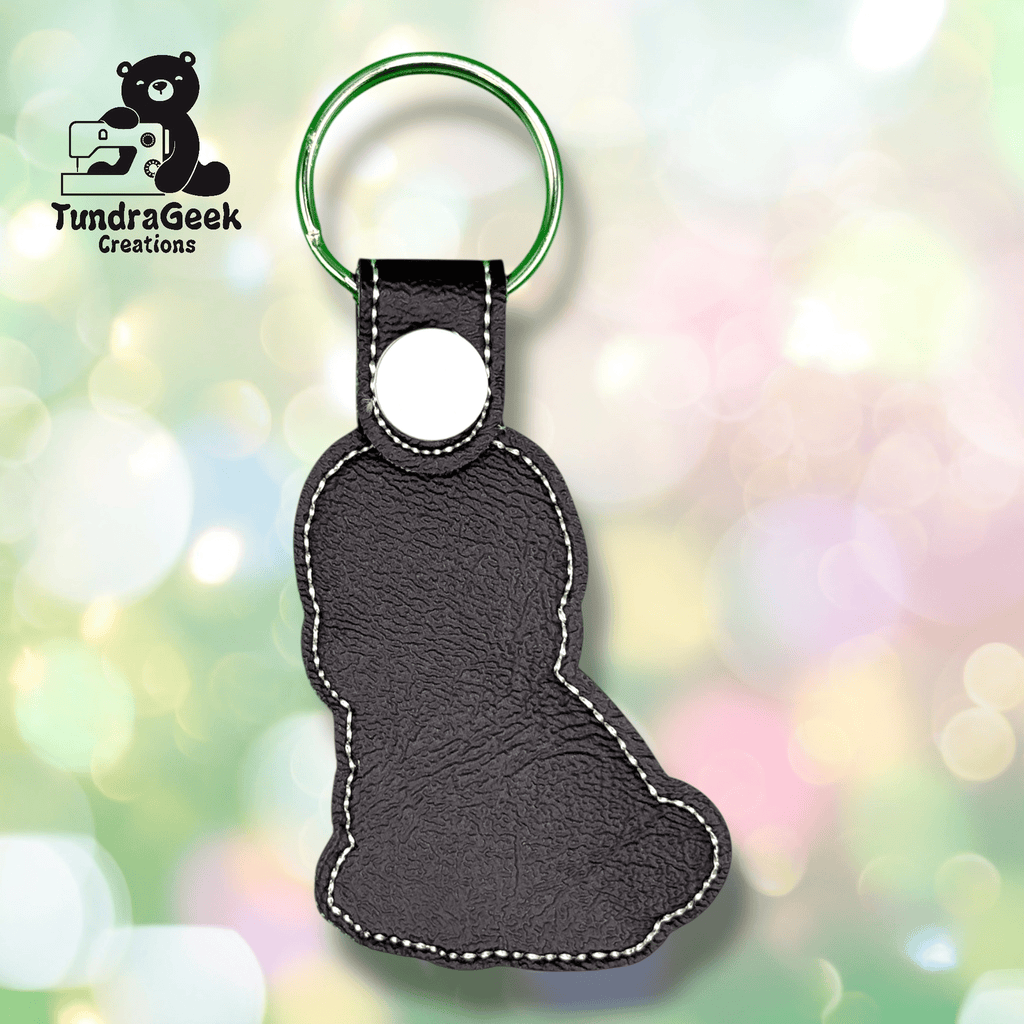 Black faux leather keychain in the shape of a sitting bear, with white stitching and a green key ring, shown on a blurred pastel background. TundraGeek Creations logo is visible in the corner.