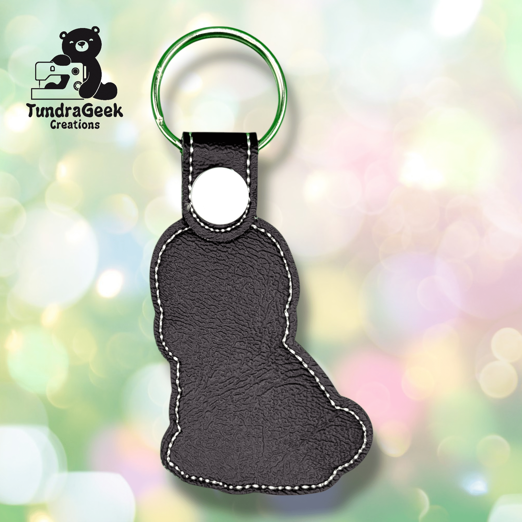 Black faux leather keychain in the shape of a sitting bear, with white stitching and a green key ring, shown on a blurred pastel background. TundraGeek Creations logo is visible in the corner.