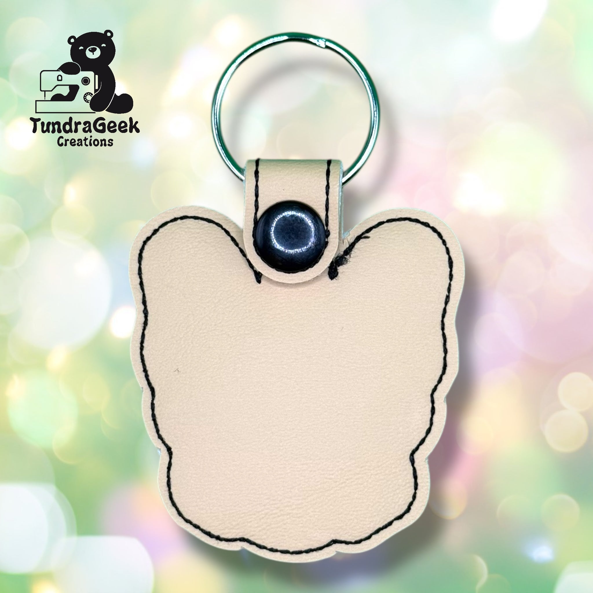 Beige, butterfly-shaped leather keychain with black stitching and a metal keyring, displayed in front of a pastel bokeh background. TundraGeek Creations logo in upper left corner.