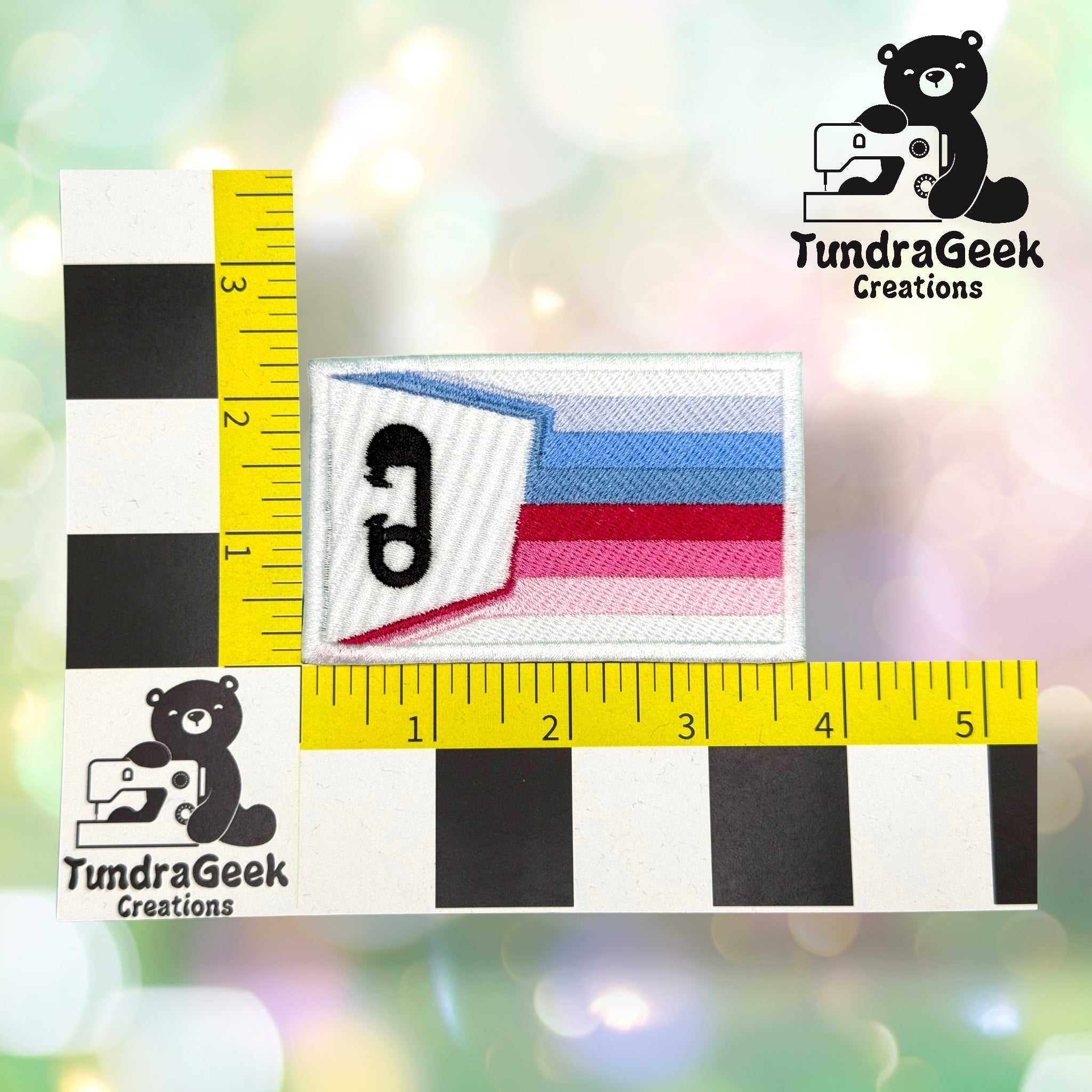 A rectangular embroidered patch with a safety pin design and blue, pink, and white stripes is shown next to a ruler for size reference, with a TundraGeek Creations logo present.