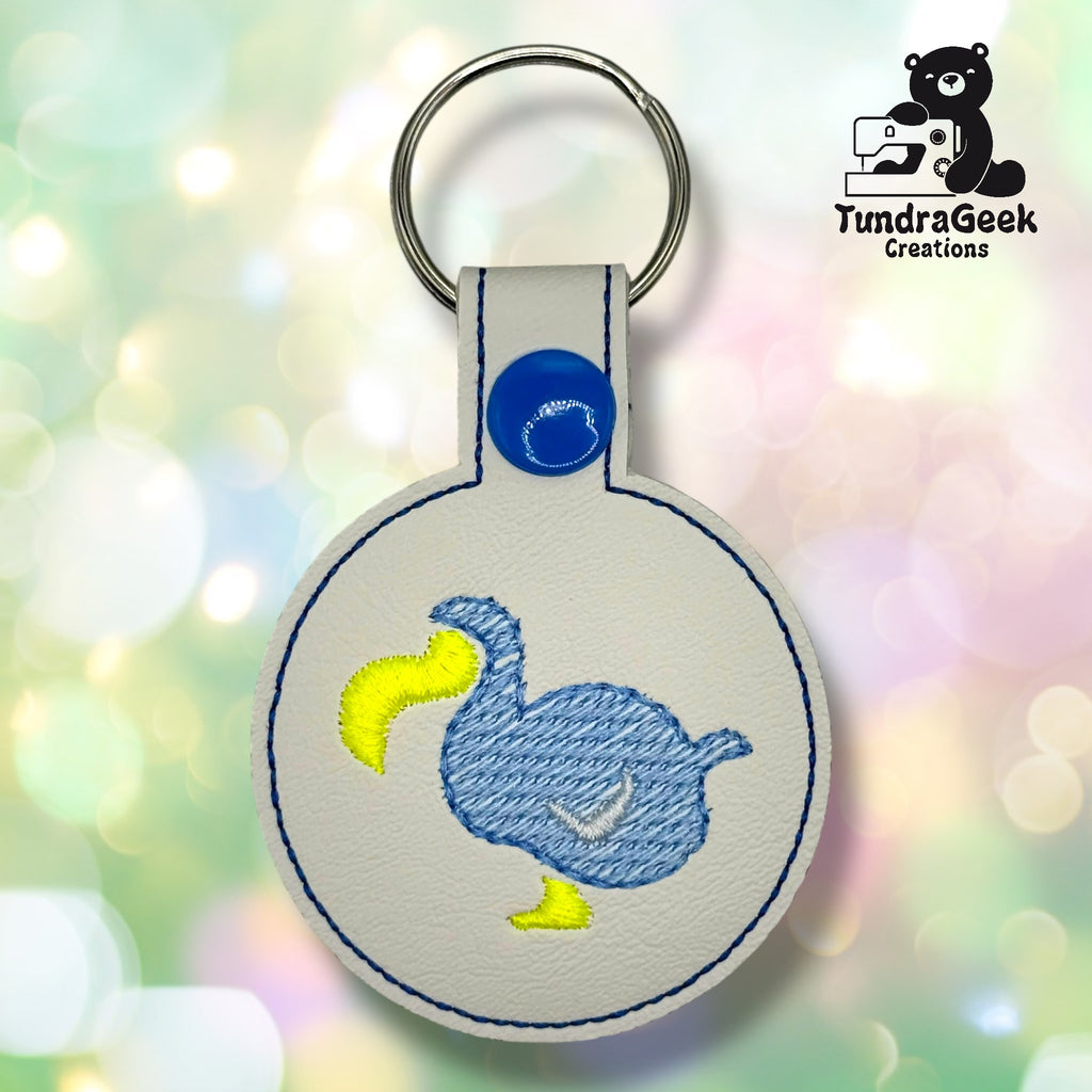Round white keychain with a blue embroidered dodo bird and yellow beak, blue stitching, and a snap closure. TundraGeek Creations logo with a bear and sewing machine in the corner.