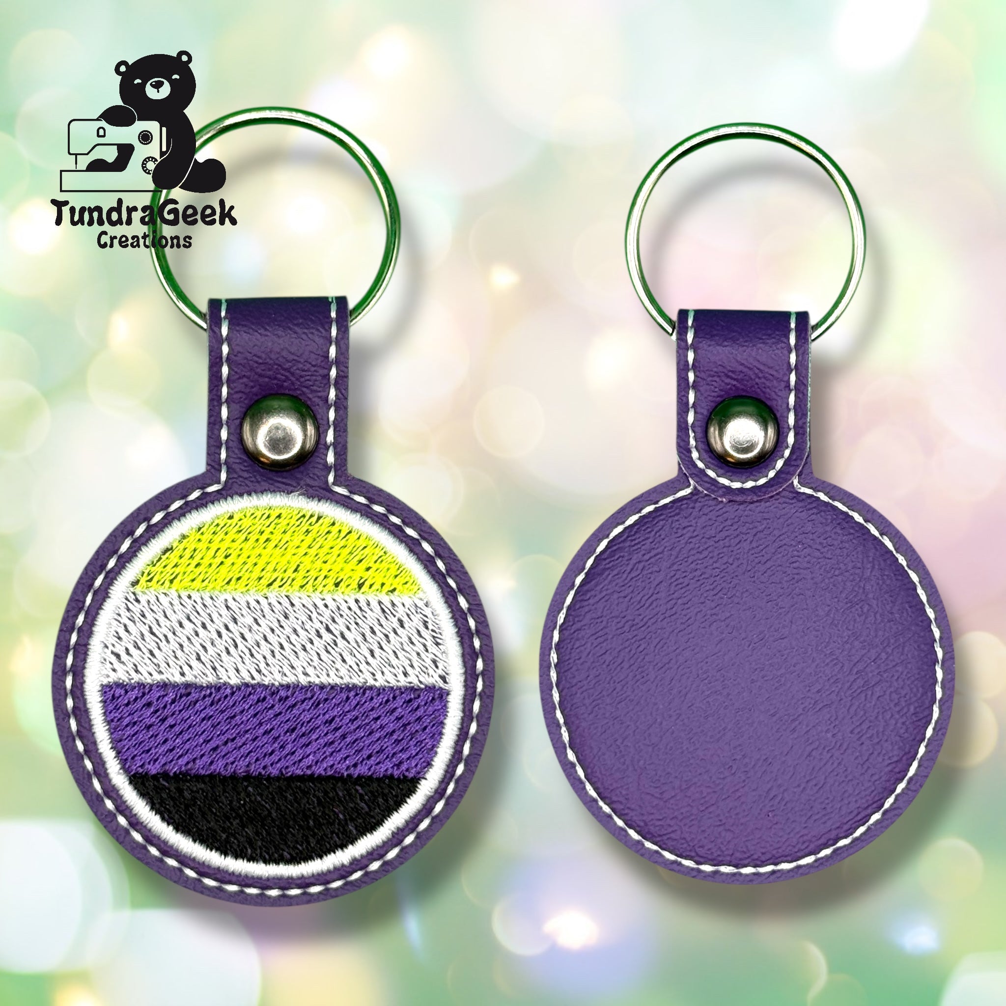 Two round purple leather keychains are shown; one displays the nonbinary pride flag, and the other shows the plain back. The Tundra Geek Creations logo appears in the top left corner.