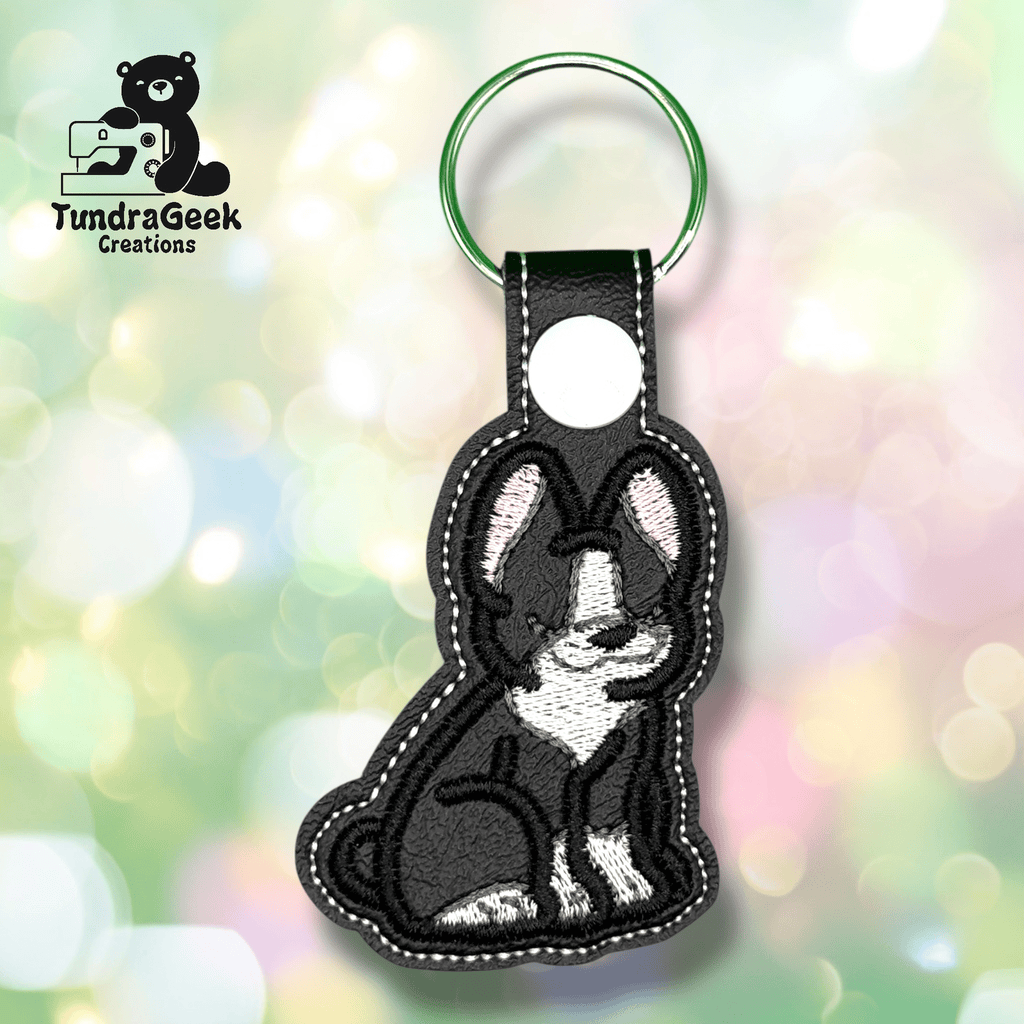 A black and white dog keychain with stitched details, attached to a green key ring, displayed against a soft, blurred pastel background. TundraGeek Creations logo is in the top left corner.