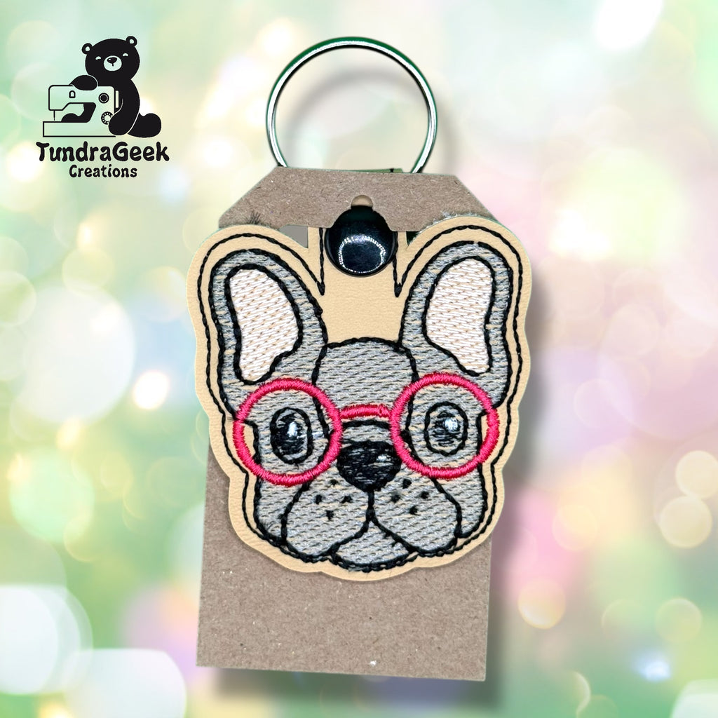 A keychain featuring an embroidered cartoon French bulldog face with red glasses, attached to a brown tag, with a TundraGeek Creations logo in the corner.