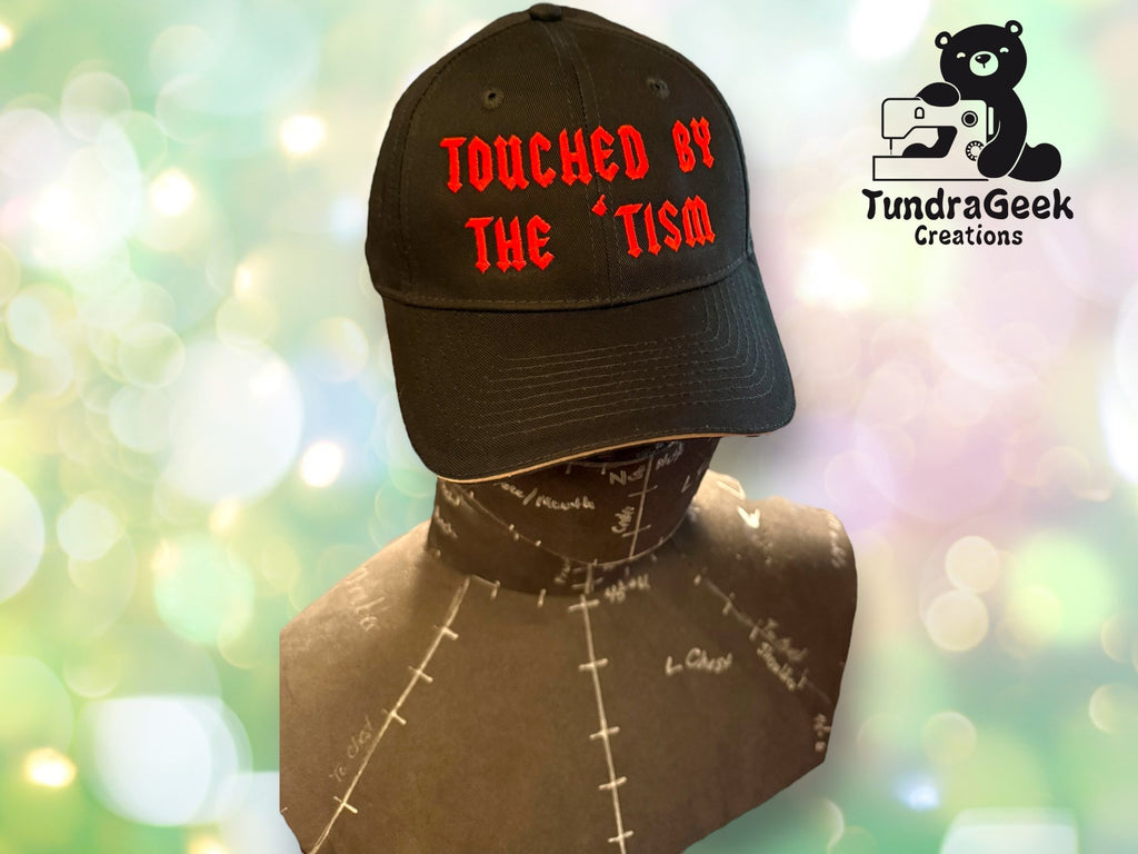A black cap with red text reading TOUCHED BY THE TISM displayed on a mannequin bust, with the TundraGeek Creations logo on the right.
