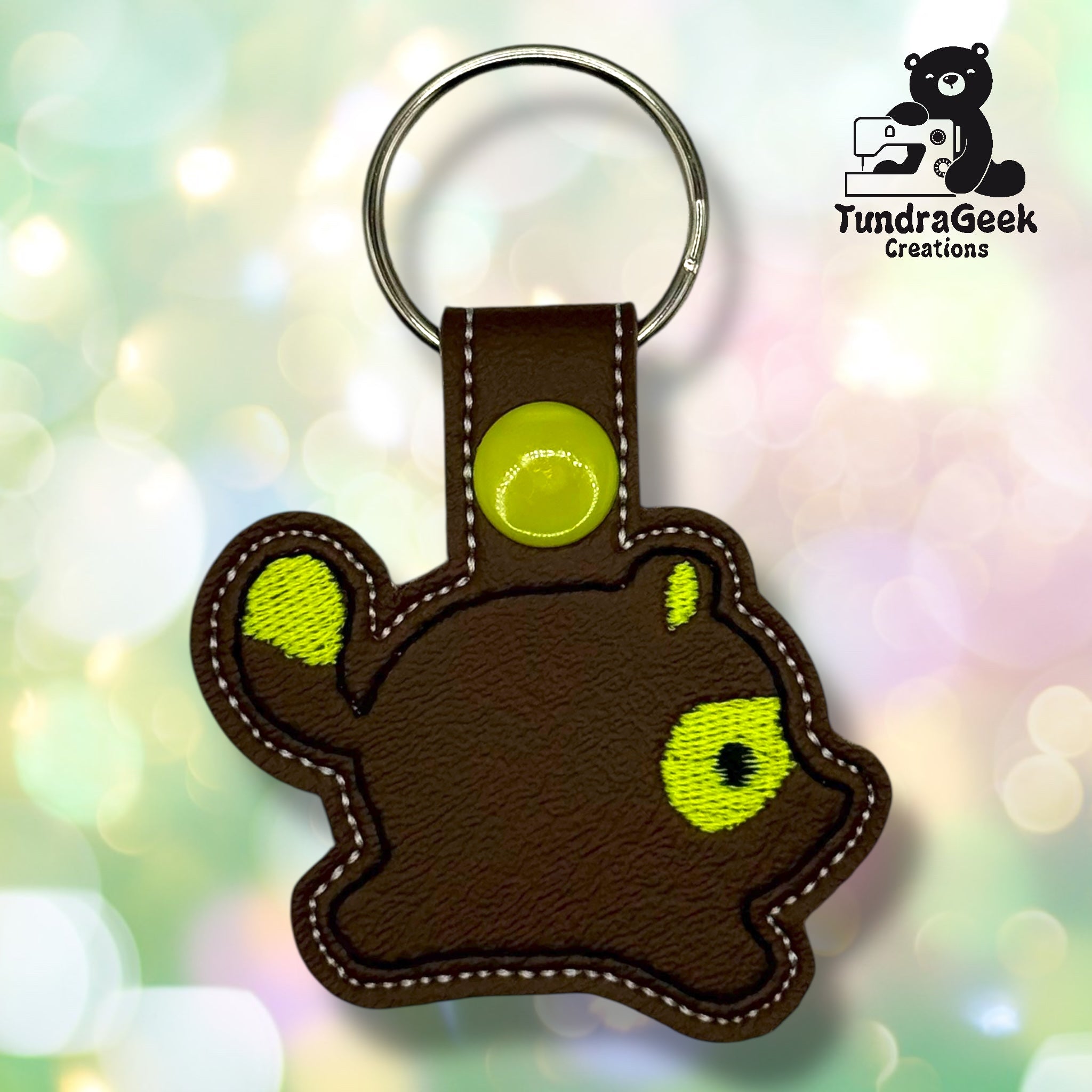 A keychain in the shape of a brown animal with a yellow eye and tail spot, made of stitched material, displayed on a blurred pastel background.