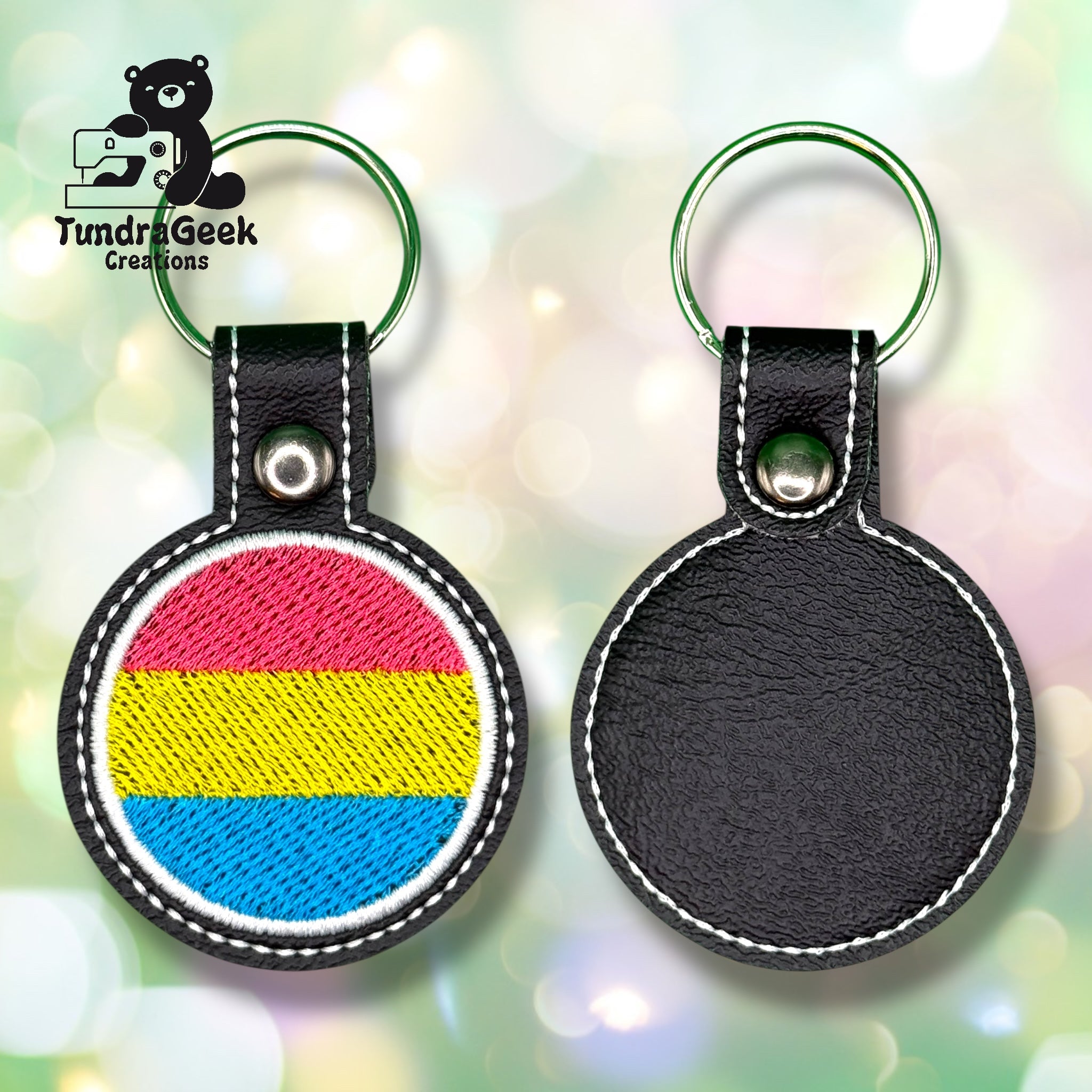 Two black leather keychains are shown; one displays the pansexual pride flag, the other is blank. Both have visible white stitching and metal rings, with a logo in the background.