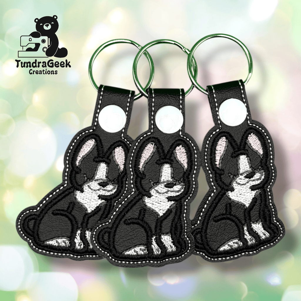 Three embroidered keychains shaped like black and white dogs with pink ears are displayed on a blurred pastel background. TundraGeek Creations logo is in the top left corner.